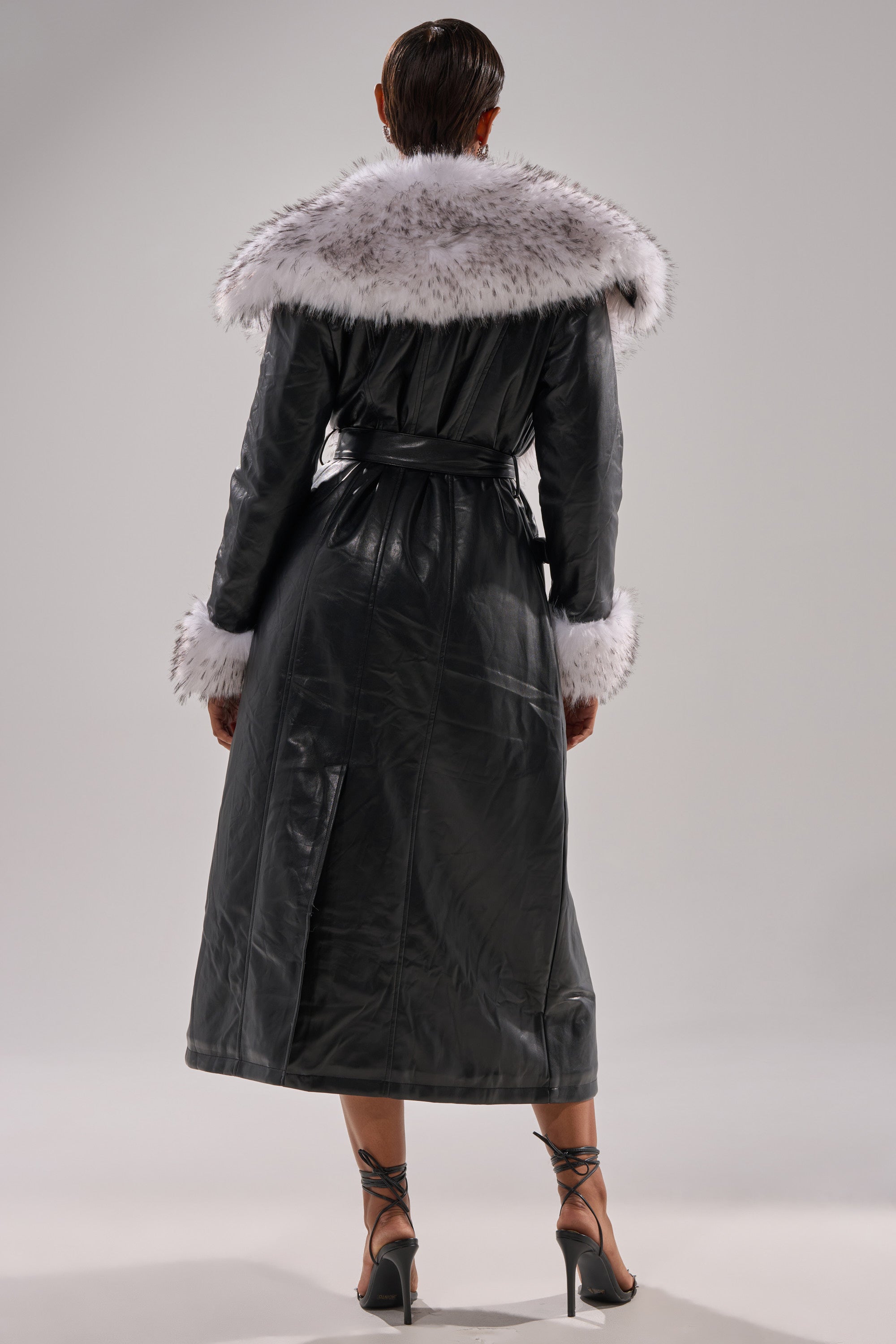 A person stands facing away, modeling the FIONA BUTTER LUXE FUR TRIM TRENCH—a long black faux leather coat with a wide white fur collar and cuffs—paired with black ankle-strap high-heeled sandals against a plain light background.