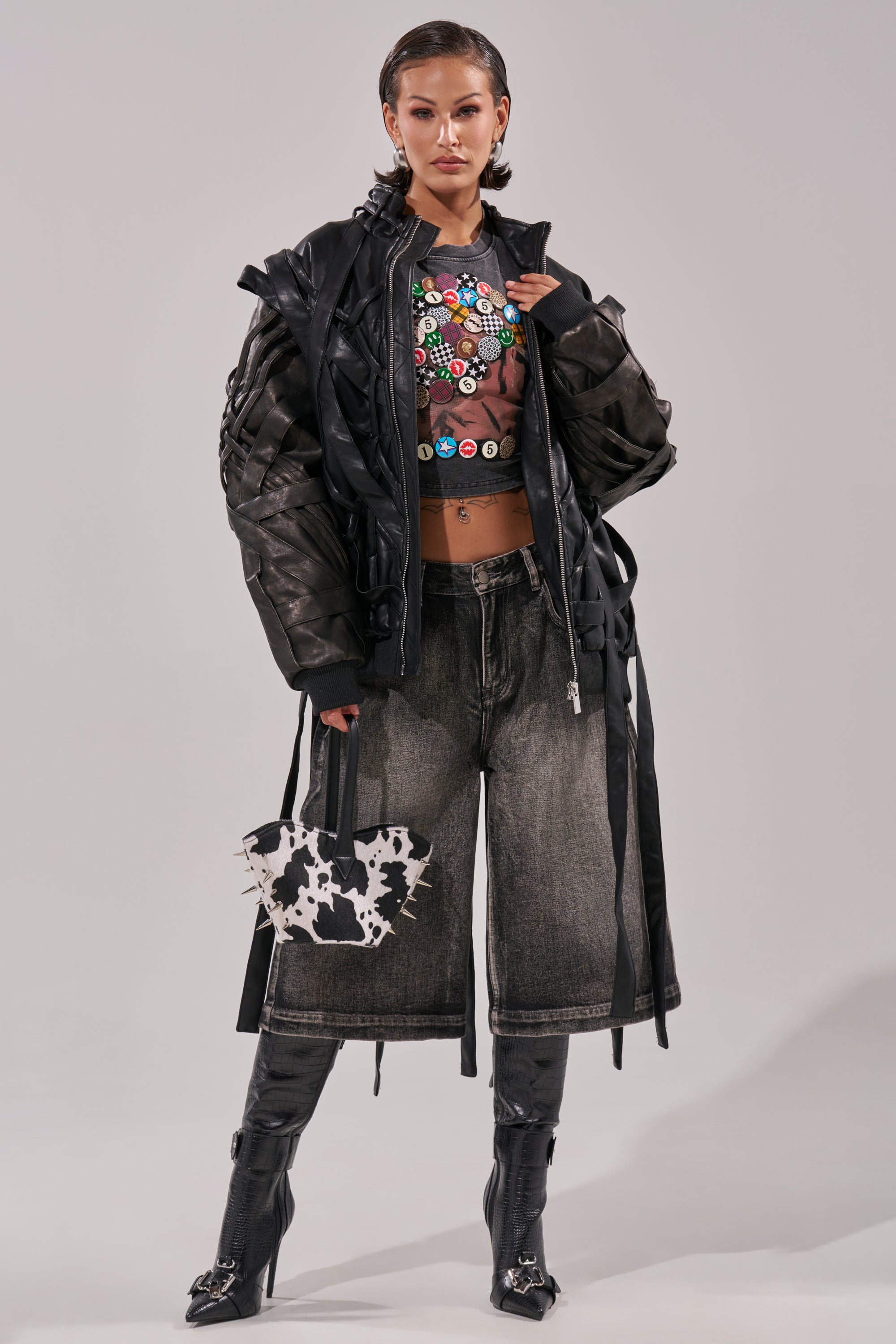 A model exudes confidence in the LOCKED IN STRAP BOMBER with quilted lining, styled over a colorful-button crop top, wide-leg faded jeans, tall black boots, and a cow-print handbag; her hair is slicked back. 9
