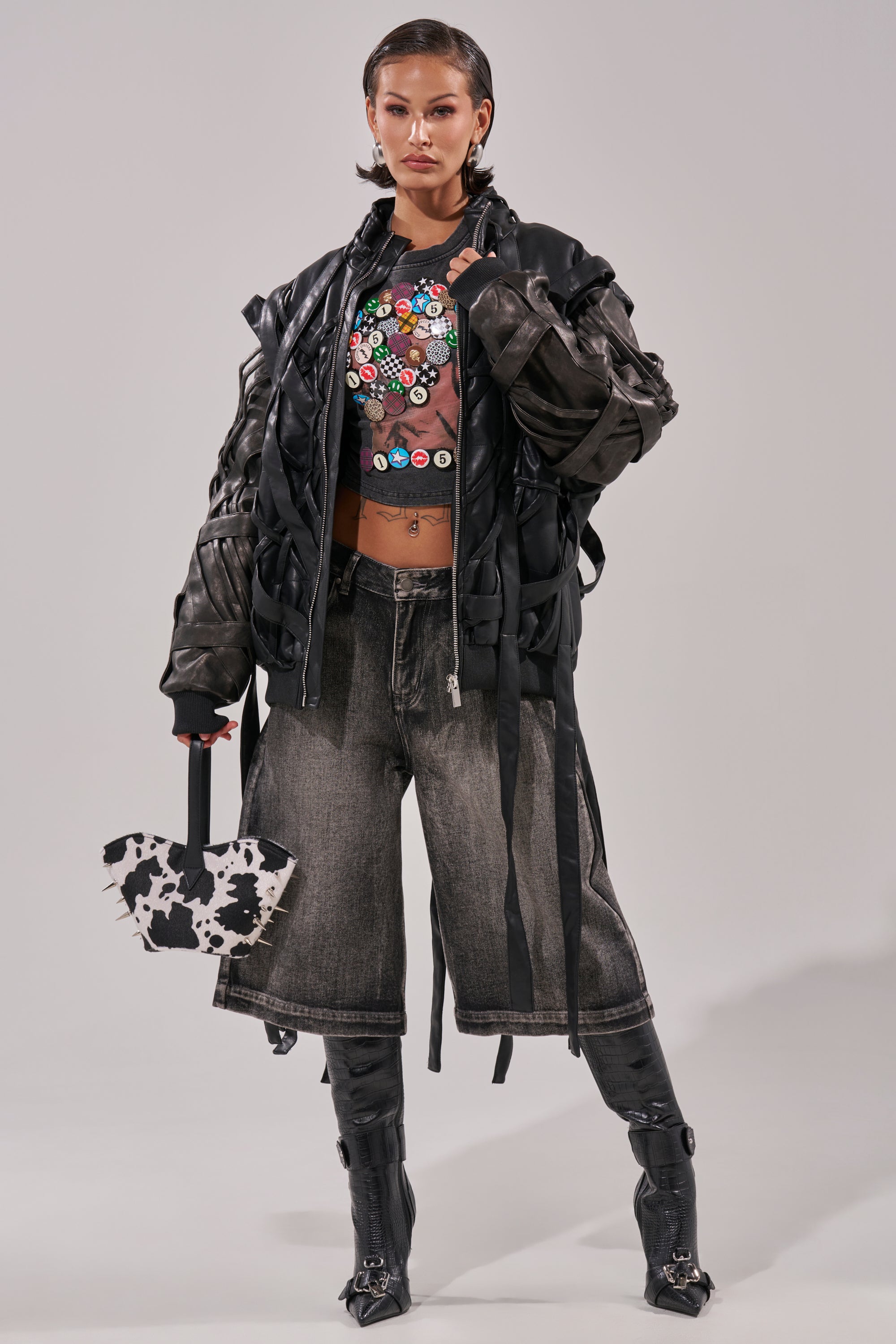 A woman stands confidently wearing the LOCKED IN STRAP BOMBER, a cropped shirt with colorful buttons, wide-leg faded jeans, tall black boots, and carrying a black-and-white patterned handbag. 10