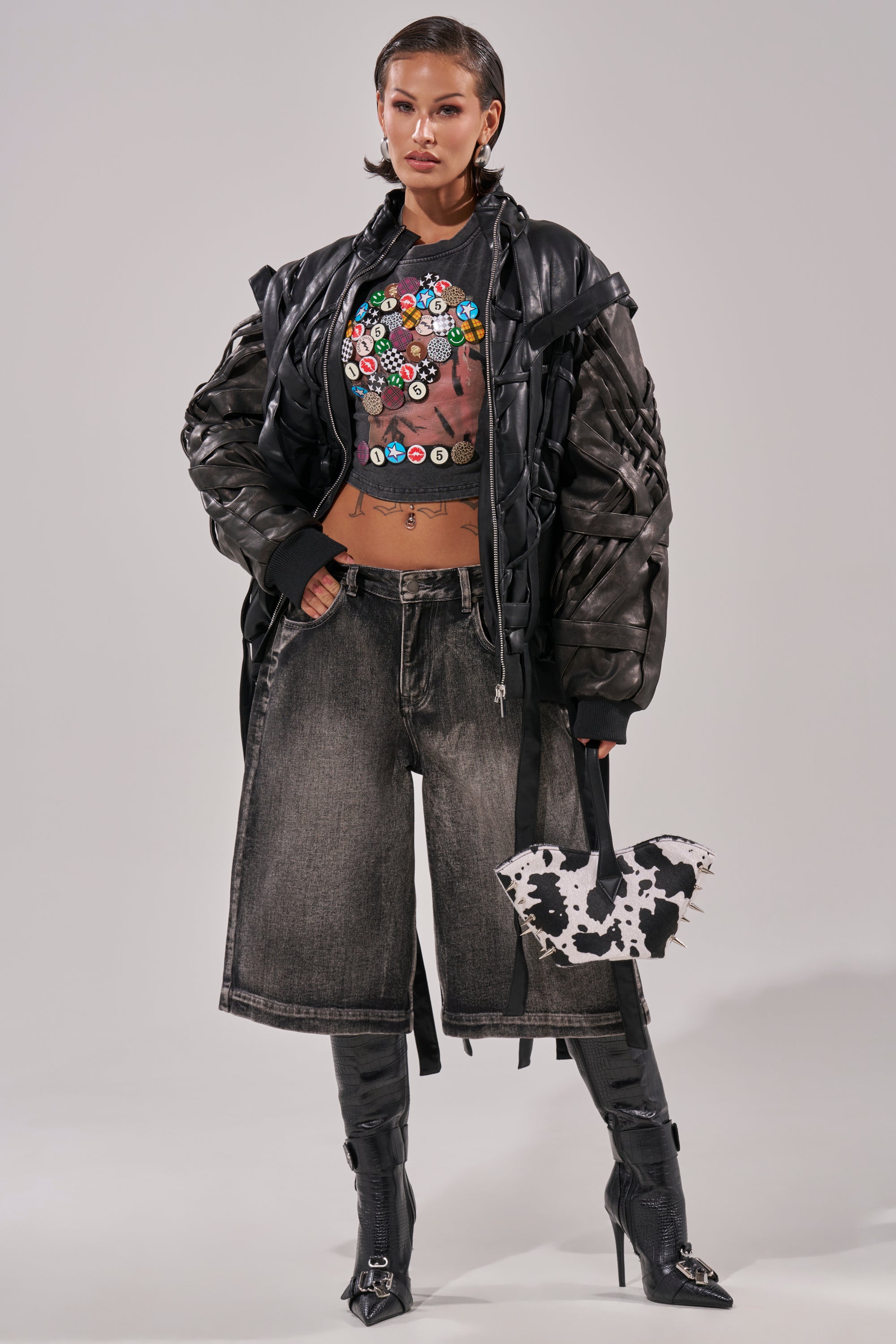 A woman stands confidently wearing the LOCKED IN STRAP BOMBER, a graphic crop top with colorful buttons, wide-leg faded shorts, black knee-high boots, and holding a cow-print handbag. 11