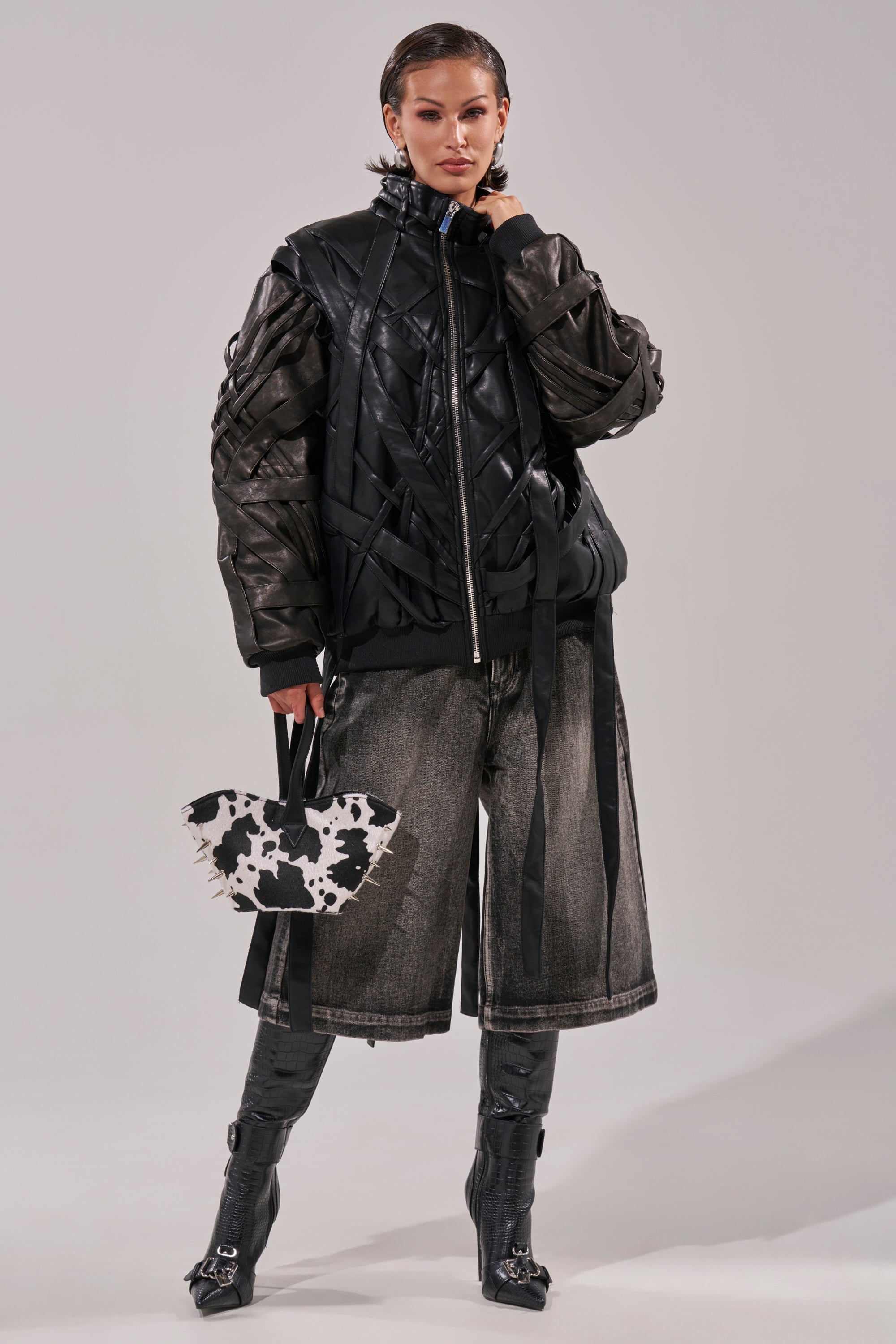 A woman poses confidently in the LOCKED IN STRAP BOMBER, paired with wide-leg faded gray denim shorts, knee-high black boots, and a small cow-print handbag. Her hair is slicked back against a plain background. 12