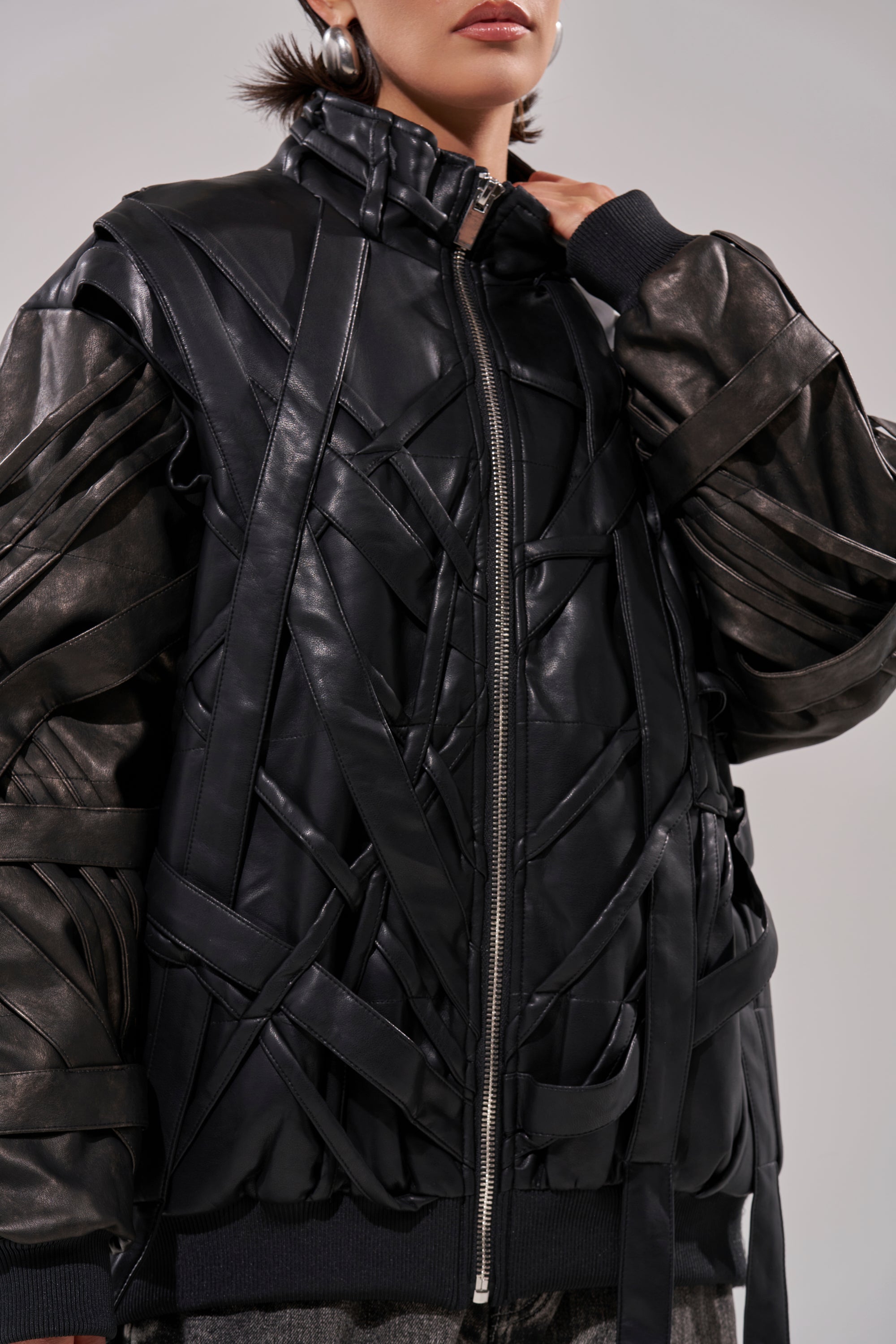 Wearing the LOCKED IN STRAP BOMBER, featuring faux leather, crisscrossing strips, and metallic zippers; only the lower half of their face is visible, with hair tucked behind their ears. 13