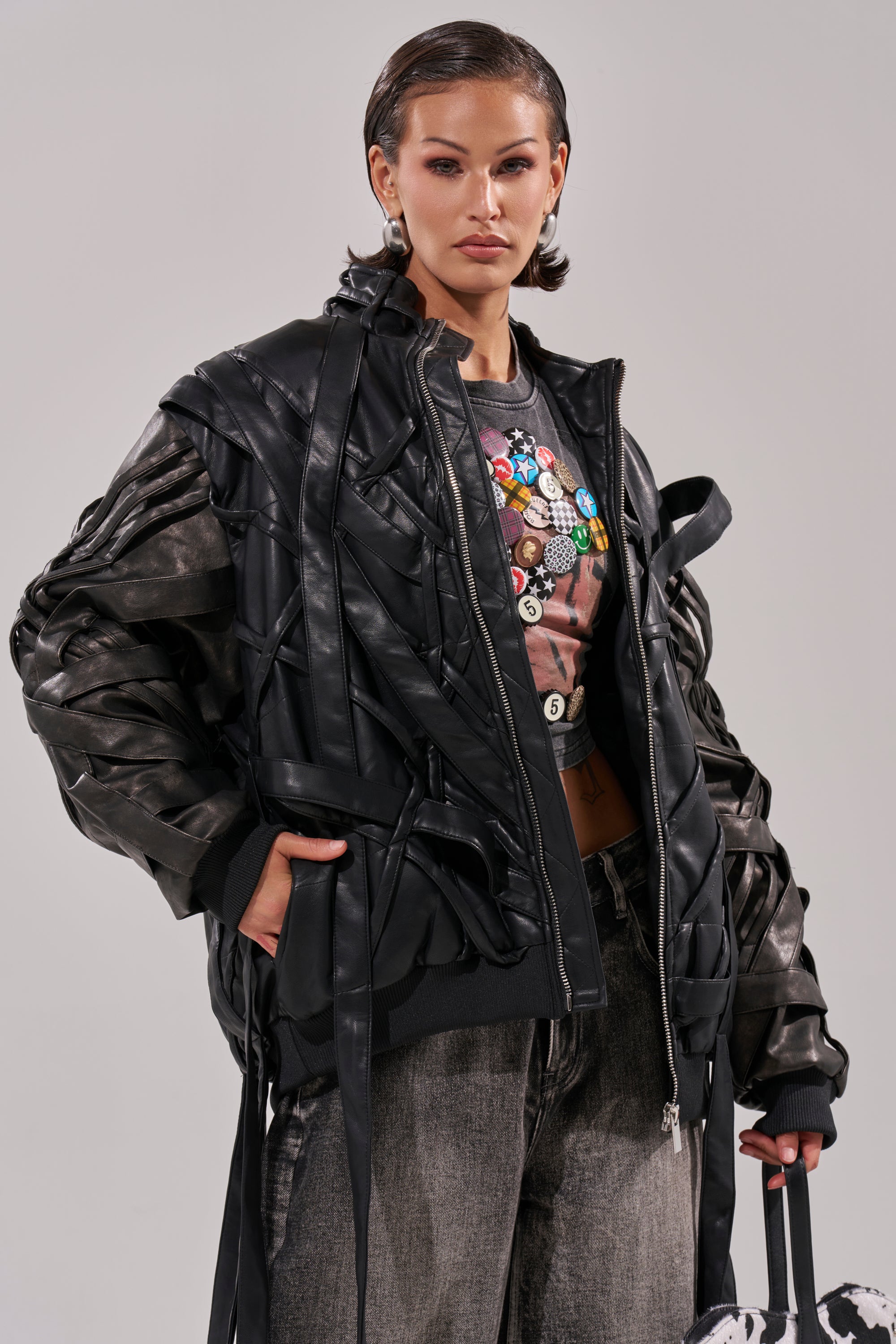 A woman with short slicked-back hair wears the LOCKED IN STRAP BOMBER over a graphic tee and gray jeans, holding a black-and-white patterned bag while posing confidently against a plain light background. 1