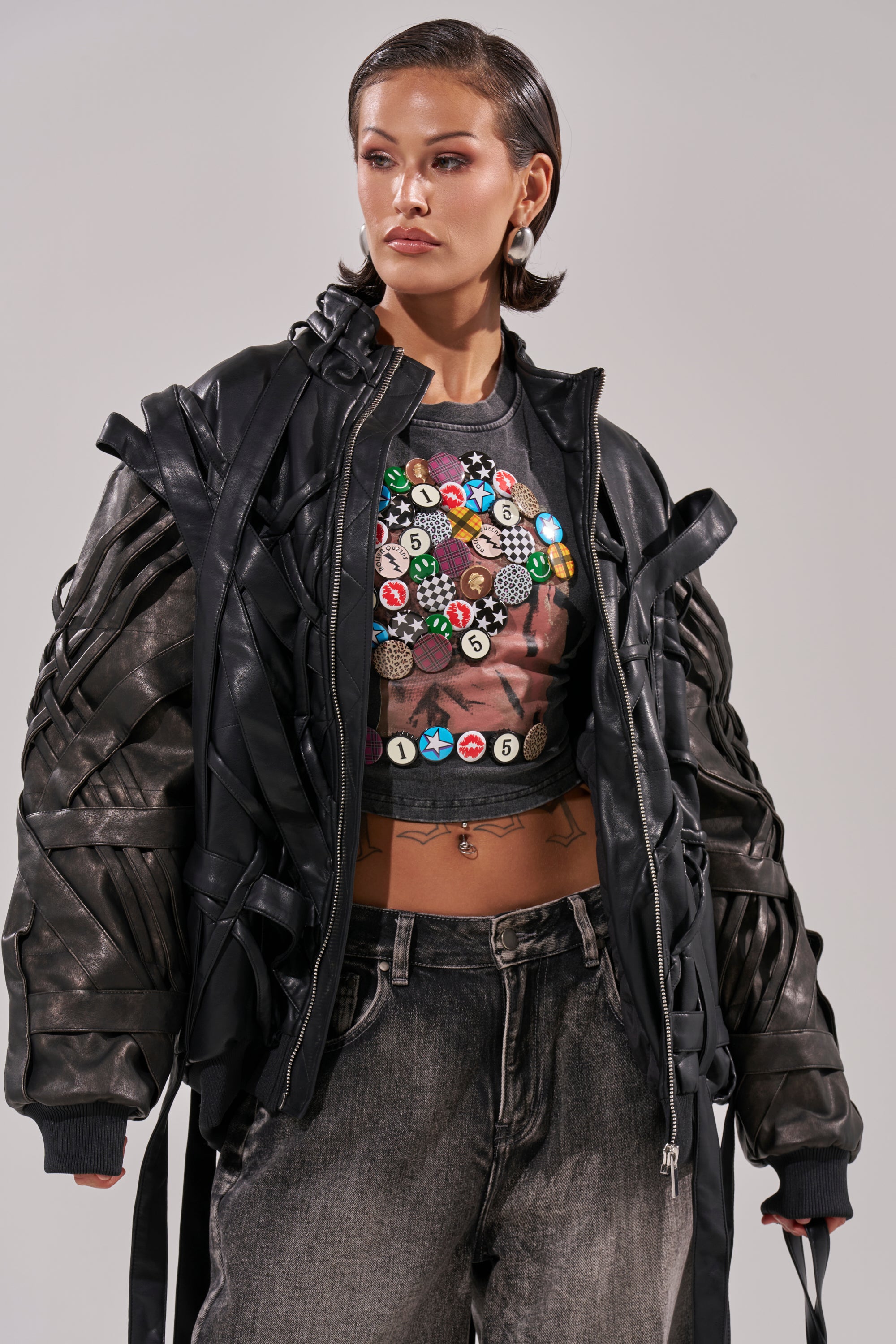 A woman with slicked-back hair wears the LOCKED IN STRAP BOMBER—an oversized jacket with quilted lining—over a crop top adorned with colorful pins and faded black jeans, standing confidently against a plain background. 2