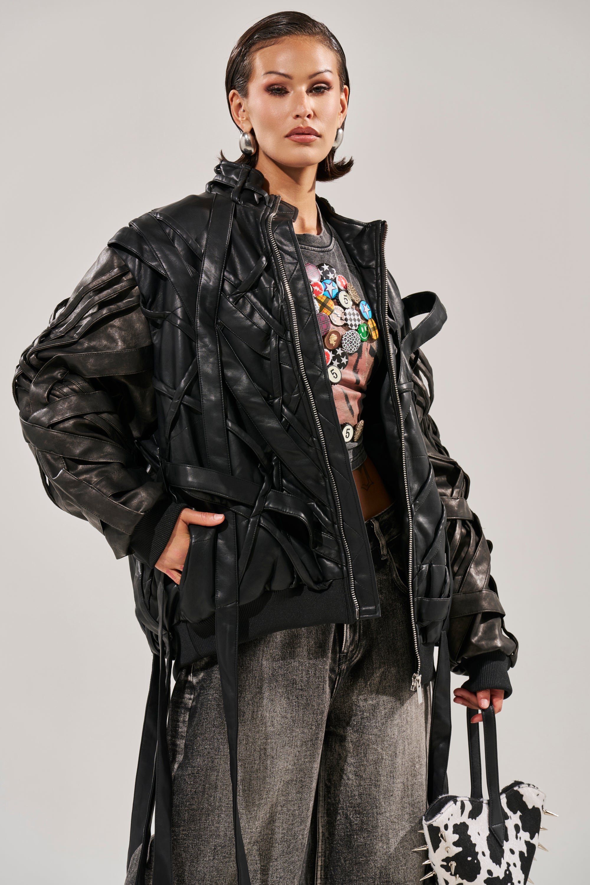 A stylish woman wears the LOCKED IN STRAP BOMBER, an oversized faux leather jacket with ribbon details, over a graphic tee and gray pants. She accessorizes with hoop earrings and a cow print bag, posing confidently against a plain background. 3