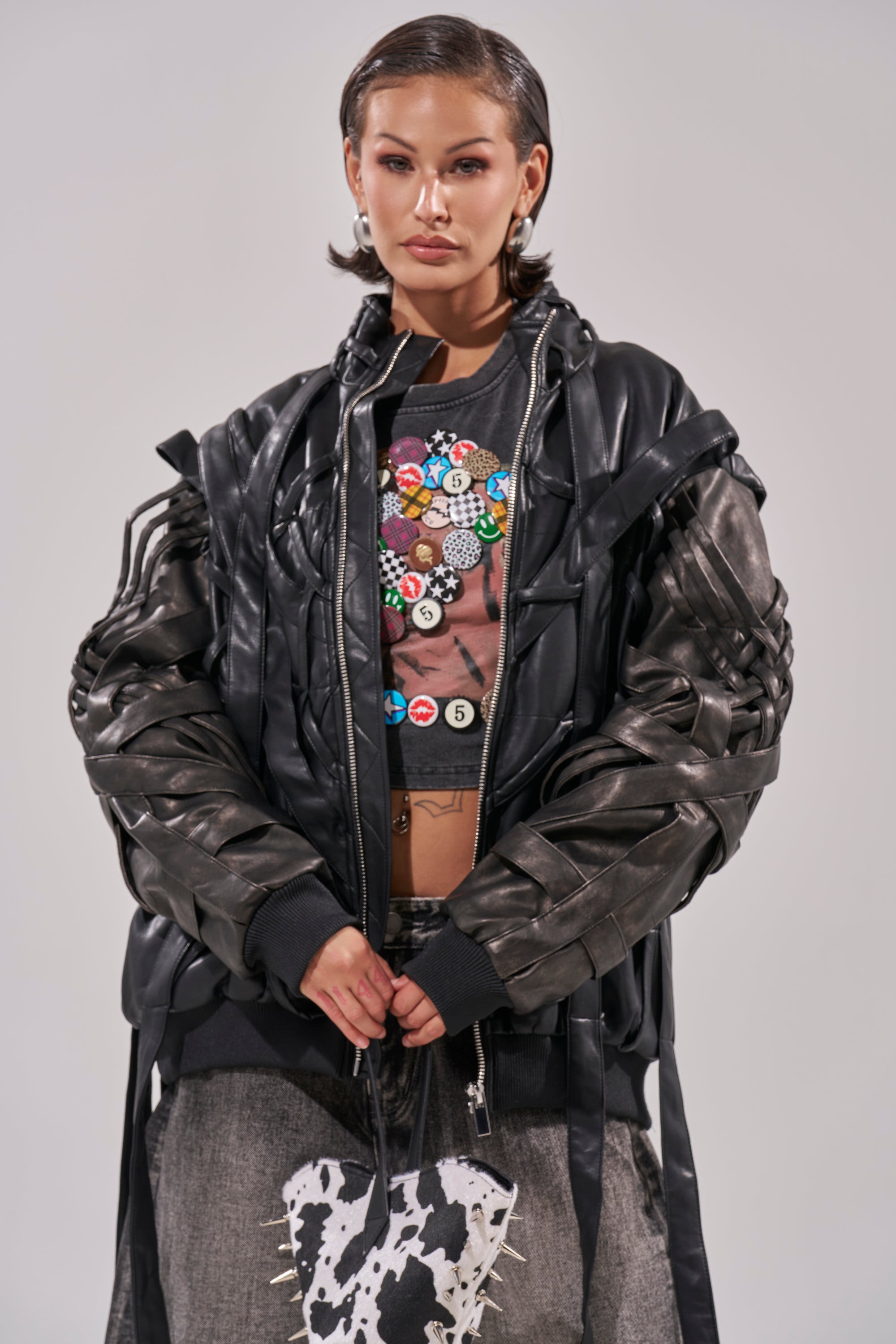 A woman with slicked-back hair wears the LOCKED IN STRAP BOMBER—a layered black faux leather jacket in an oversized fit—with a brown crop top featuring colorful buttons, holding a black-and-white cow-print bag against a plain background. 4