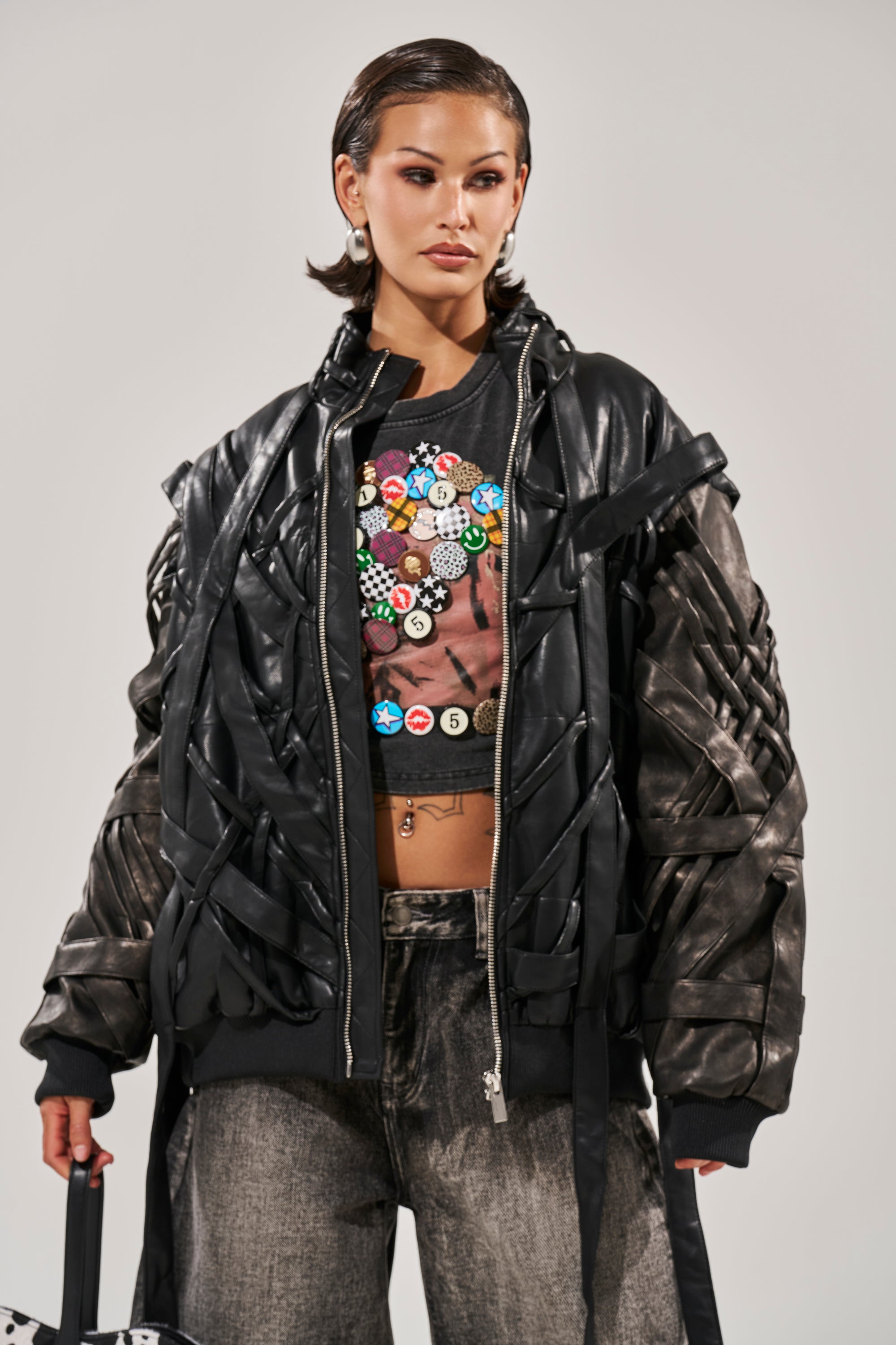 A woman poses confidently in the LOCKED IN STRAP BOMBER, a sculptural oversized black leather jacket with woven details, paired with a black T-shirt with colorful buttons and high-waisted gray jeans against a plain background. 5