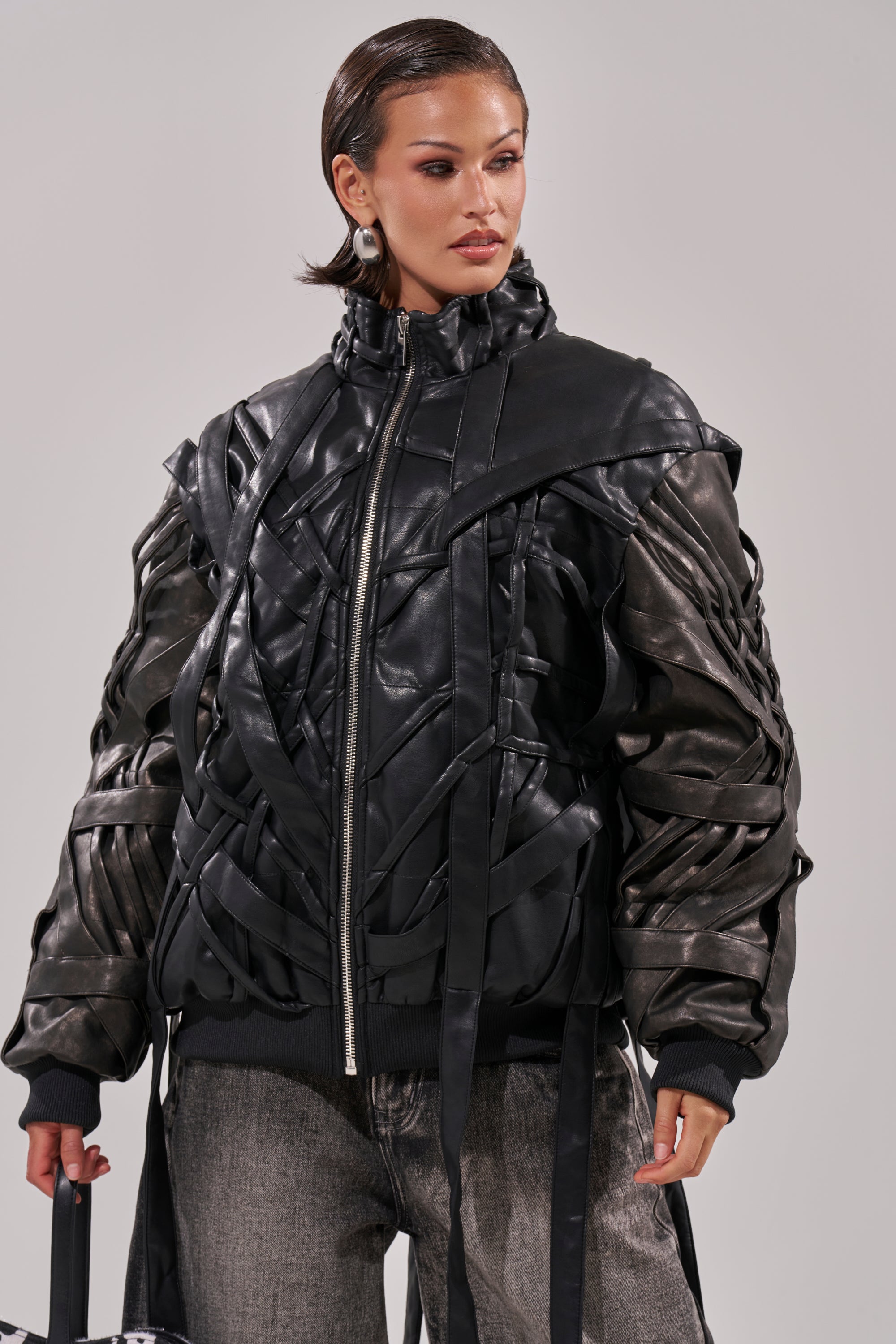 Wearing the LOCKED IN STRAP BOMBER—a textured black faux leather jacket with woven, layered details—paired with gray jeans, this person stands confidently against a plain light background, hair slicked back and gaze to the side. 6