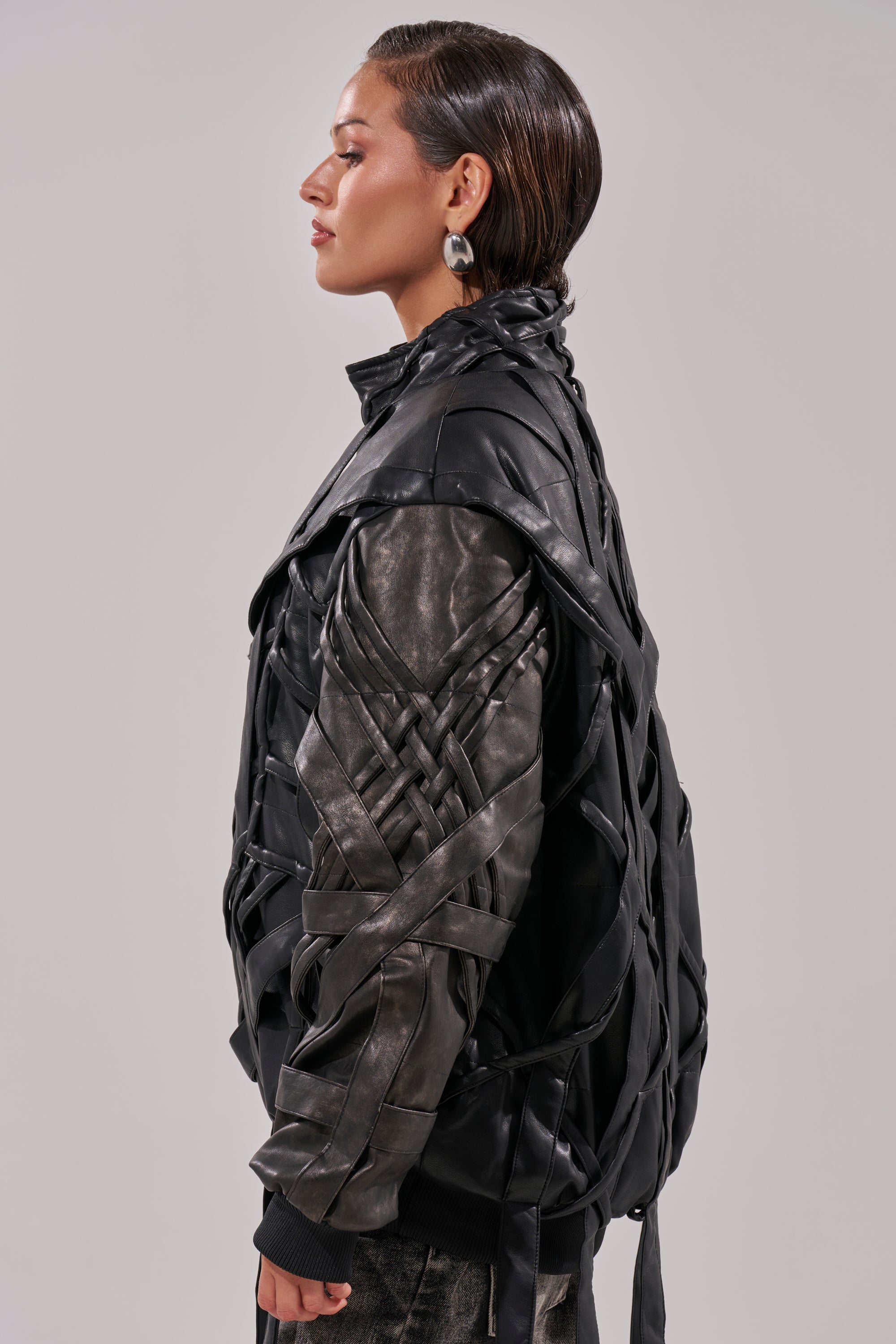 A woman in profile wears the LOCKED IN STRAP BOMBER, a black faux leather jacket with layered, crisscrossed straps. She pairs it with metallic earrings; her hair is slicked back against a plain light gray background. 7