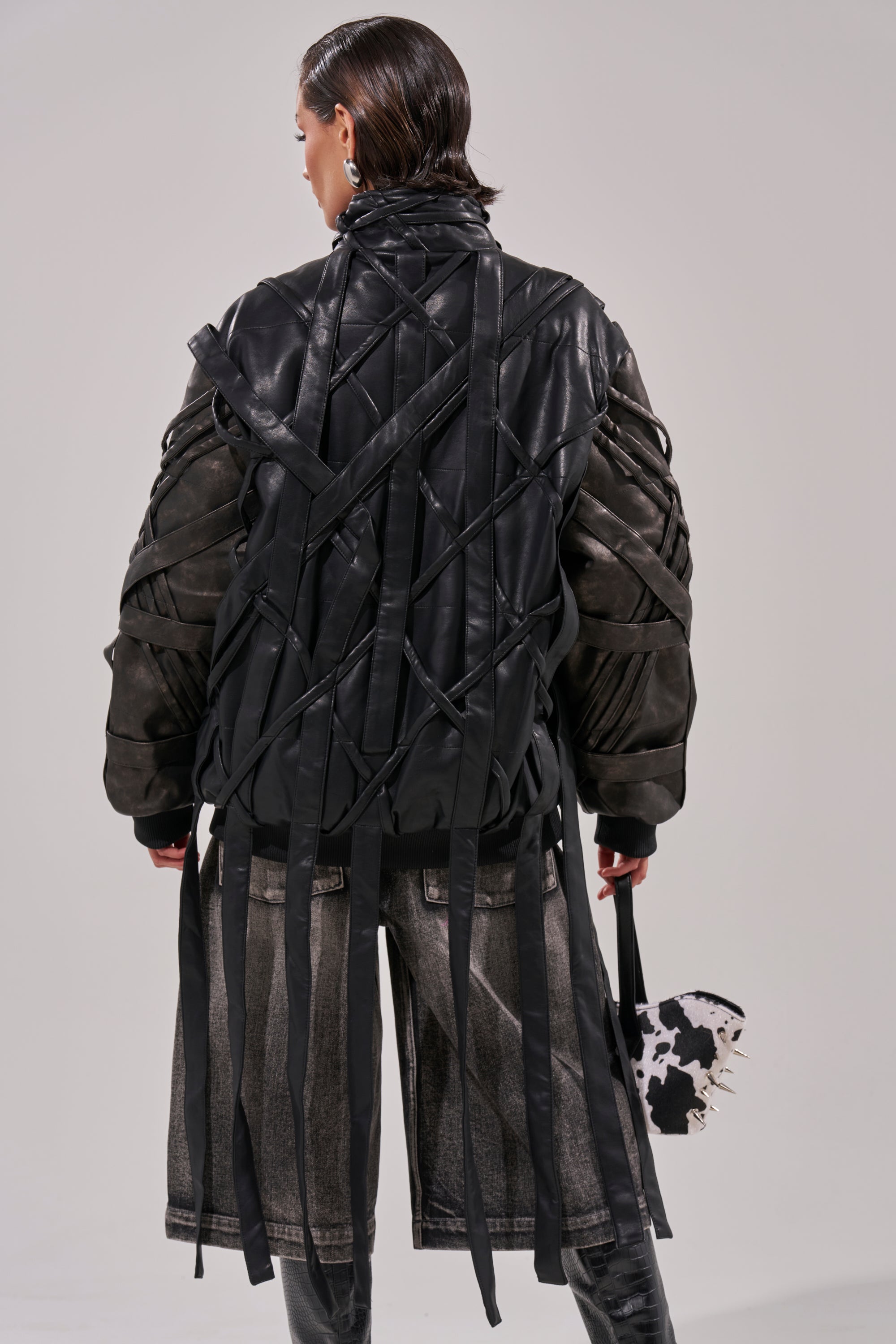 Facing away, a person wears the oversized LOCKED IN STRAP BOMBER in black and dark gray faux leather with interlaced straps and hanging ribbons, paired with loose faded shorts. They hold a small spiked cow-print bag. 8