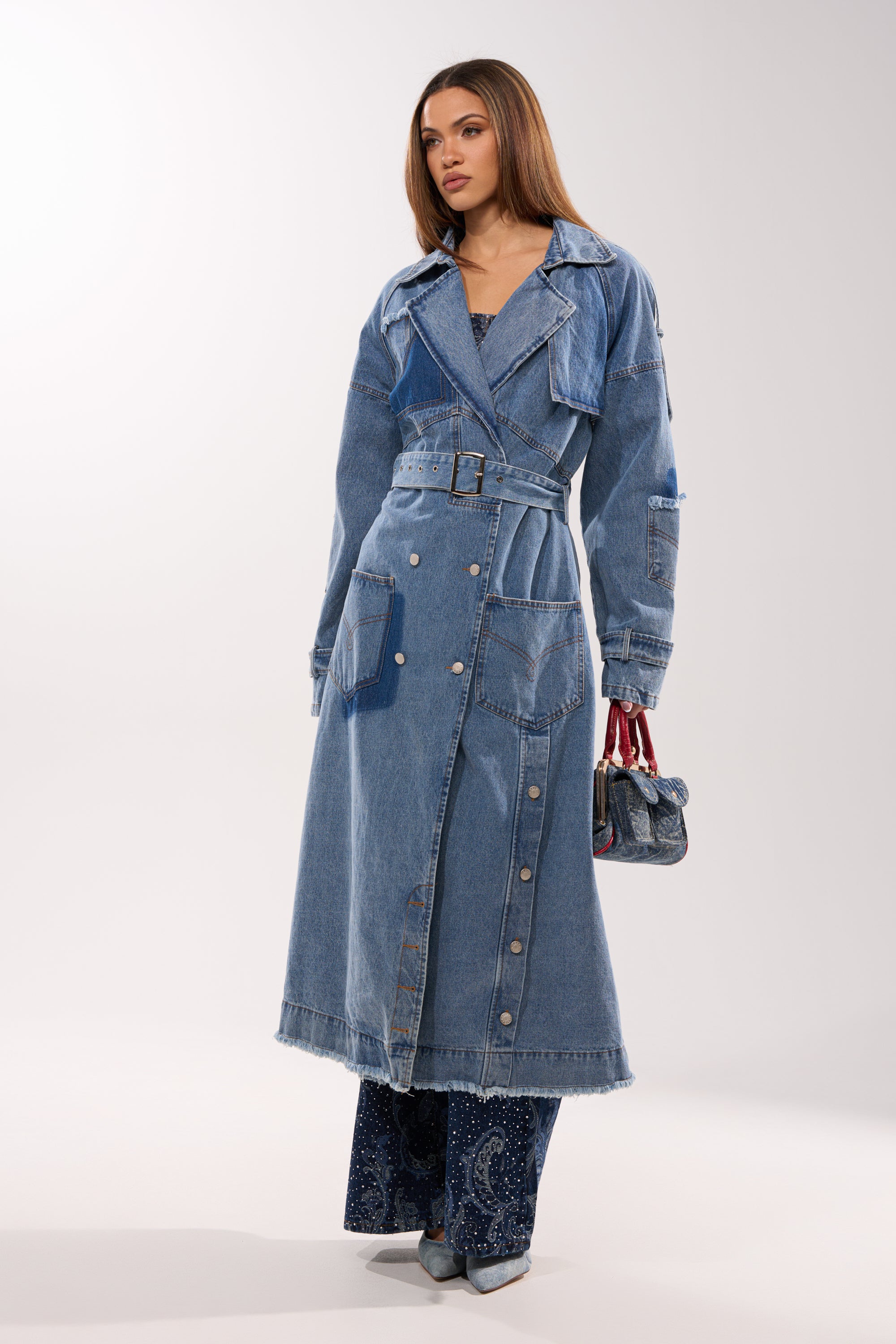 A woman wears the RILEY PATCHWORK DENIM TRENCH, a vintage-wash longline coat with silver buttons, styled over blue patterned pants. She carries a small handbag and pointed shoes, posing against a plain white background. 0