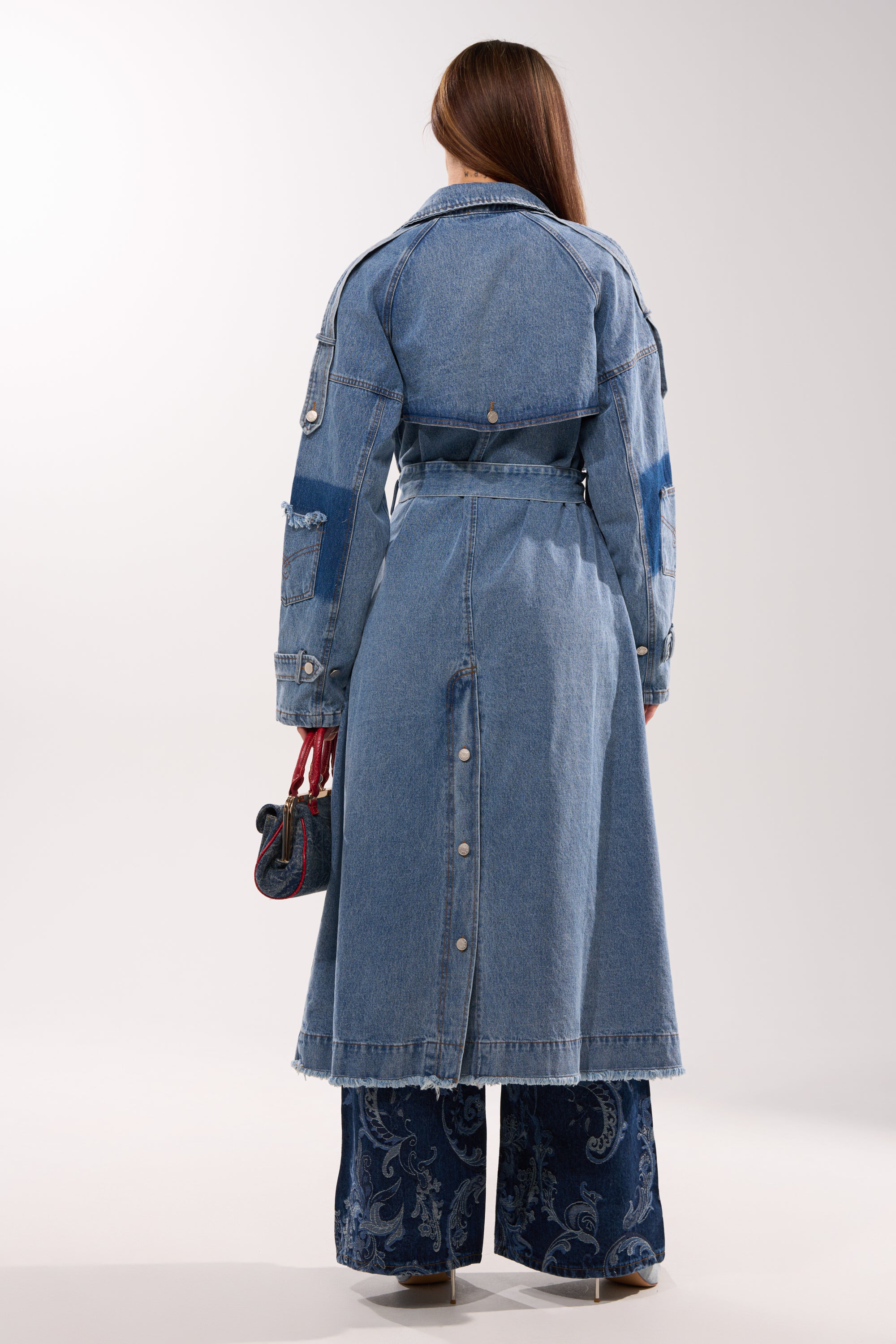 A woman with long brown hair, seen from behind, wears the RILEY PATCHWORK DENIM TRENCH over wide-leg patterned blue jeans, holding a small handbag against a plain light background. 9