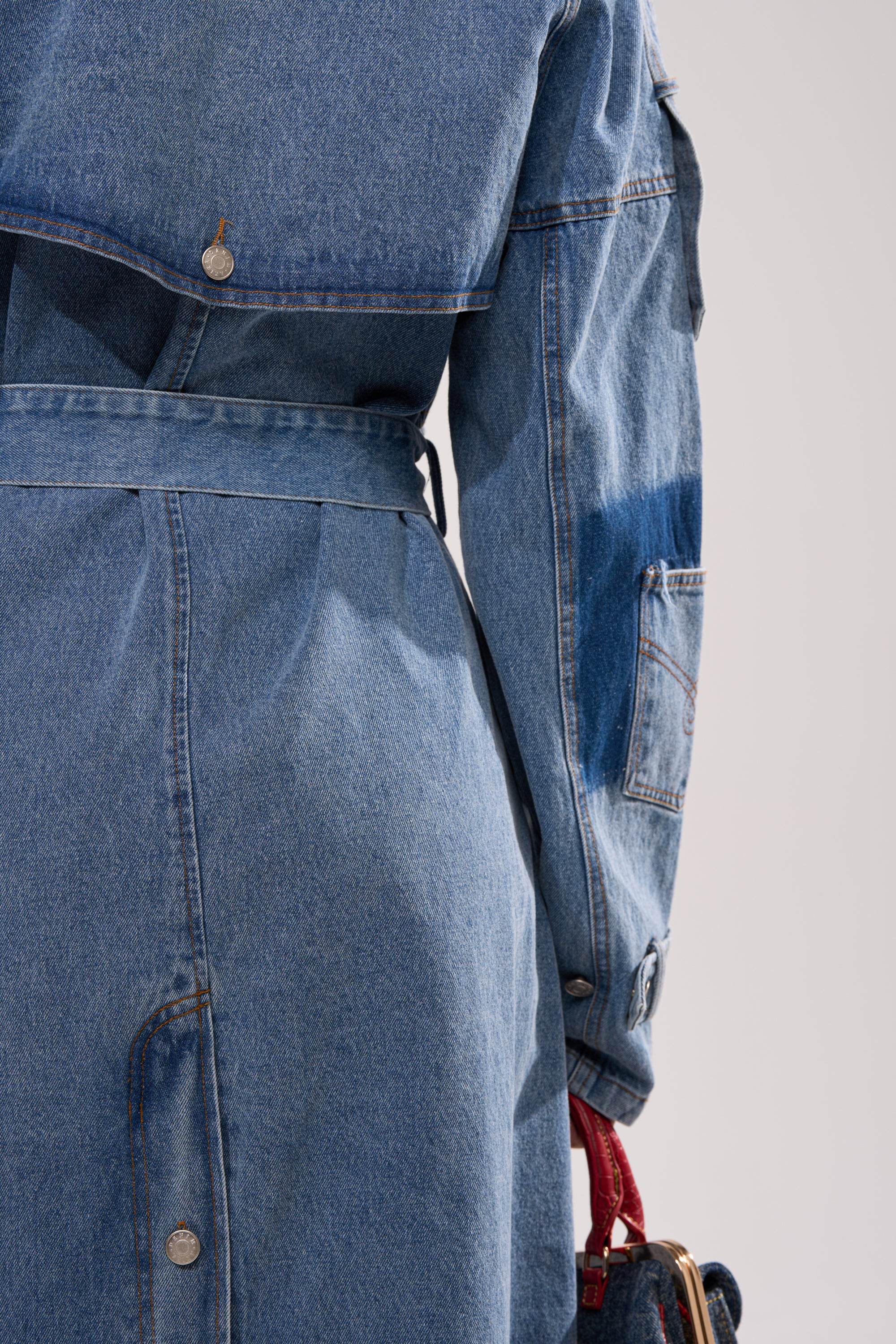 A close-up of a person wearing the RILEY PATCHWORK DENIM TRENCH, showcasing its longline silhouette and detailed back and sleeves. They hold a small denim bag with a red strap. 10