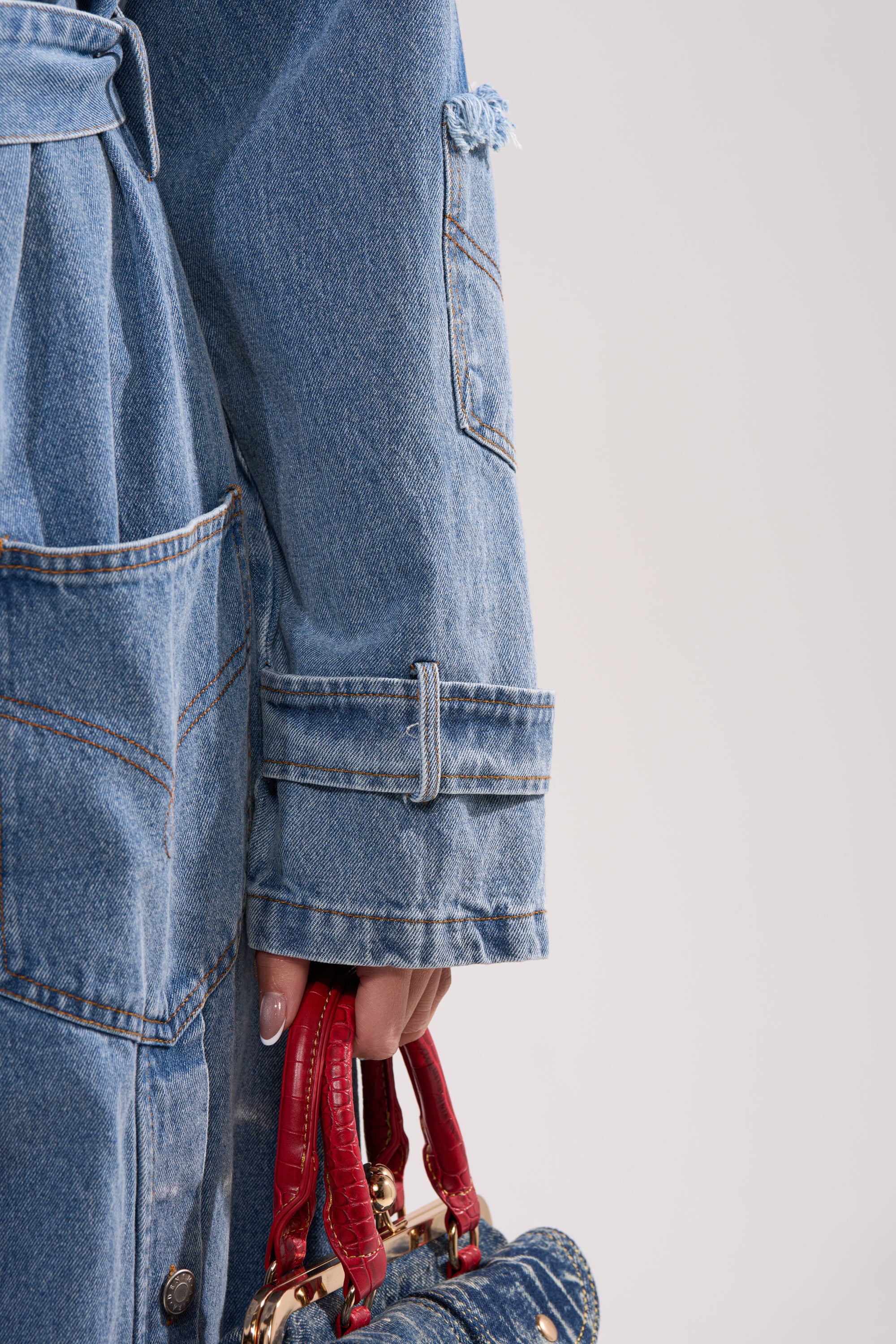 Wearing the RILEY PATCHWORK DENIM TRENCH, a person holds a small denim handbag with red handles against a light background. 12