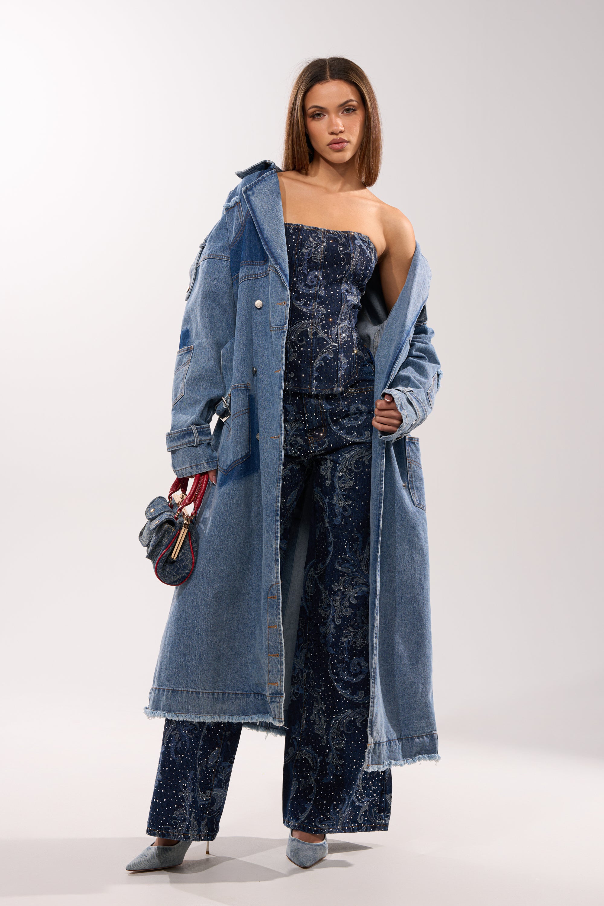 A woman stands confidently in a strapless navy blue paisley jumpsuit, gray heels, a small dark blue handbag with red handles, and the RILEY PATCHWORK DENIM TRENCH draped over her shoulders. 13