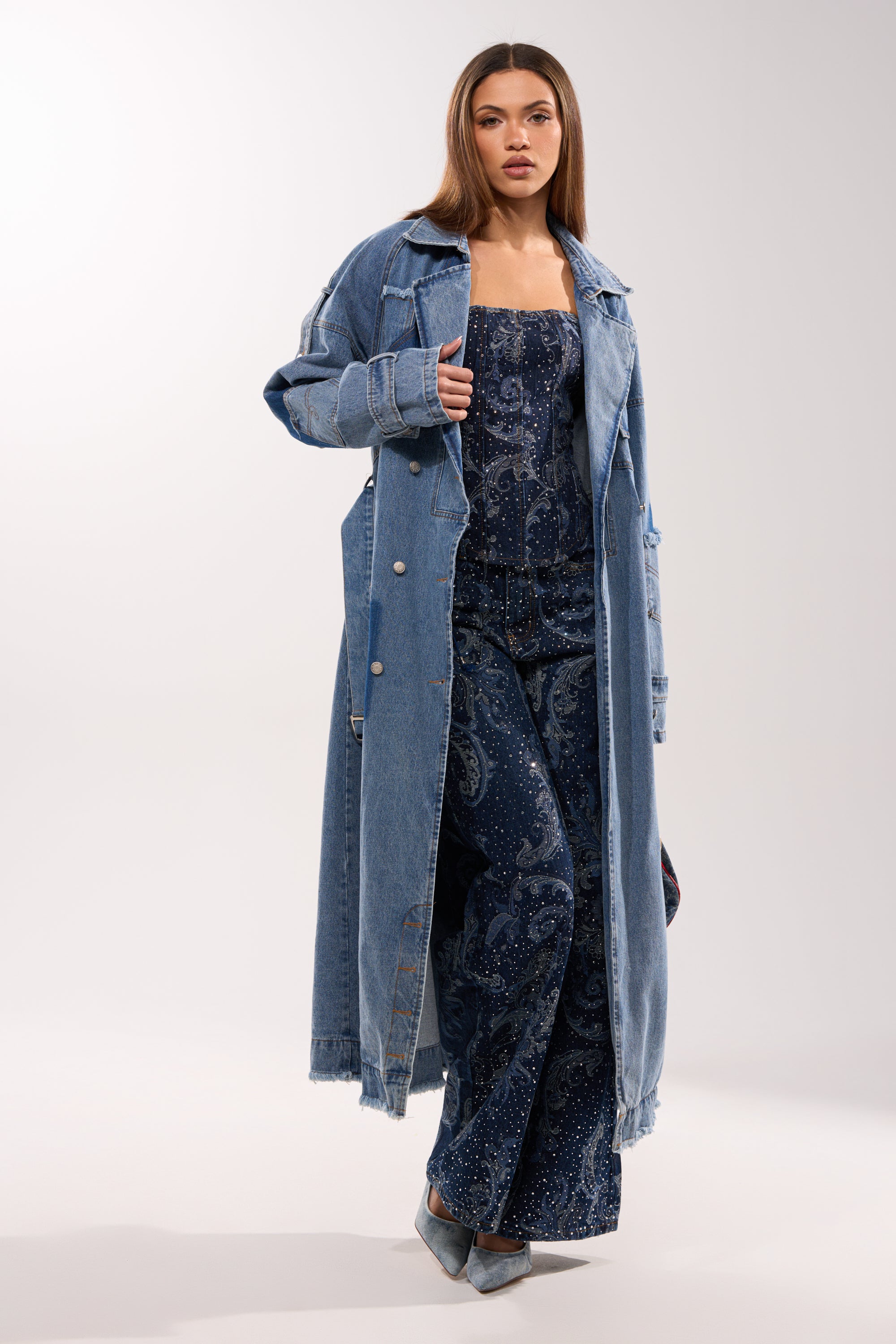A woman poses against a plain light background wearing the RILEY PATCHWORK DENIM TRENCH over a navy paisley jumpsuit and pointed-toe heels. 1