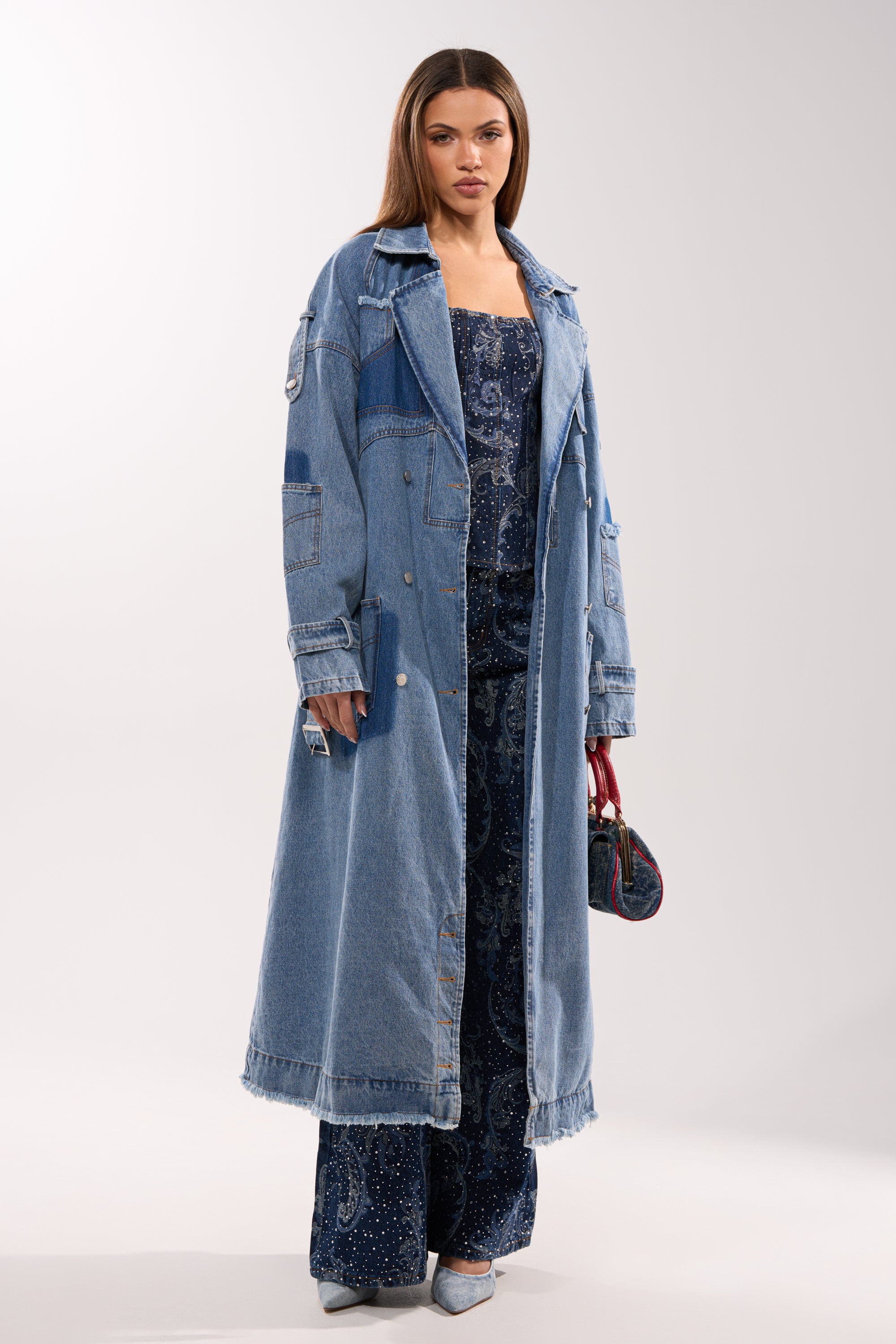 A woman stands against a plain light background wearing the RILEY PATCHWORK DENIM TRENCH over a dark blue patterned jumpsuit, accessorized with a small handbag and silver heels. 2