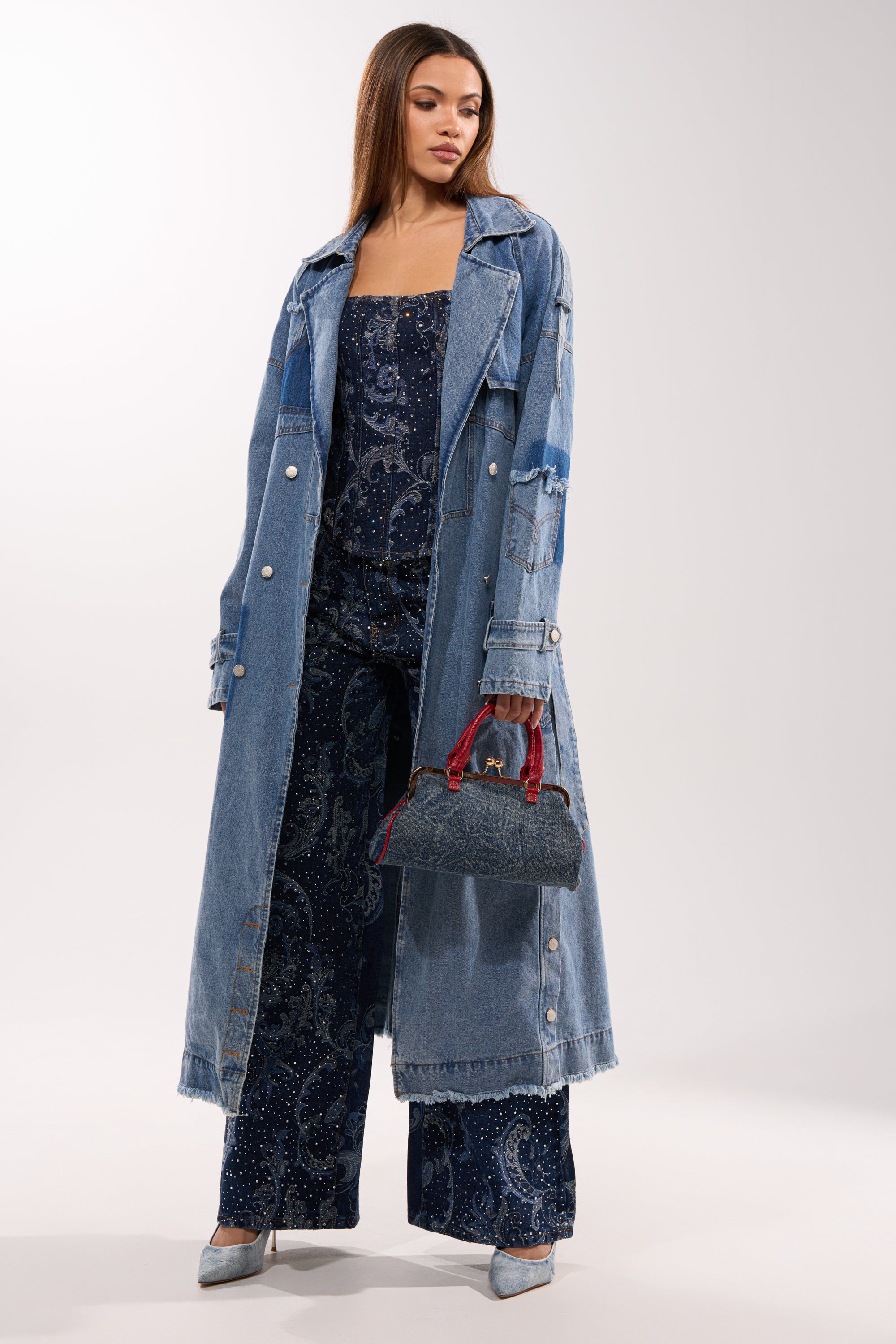 A woman in the RILEY PATCHWORK DENIM TRENCH with a longline silhouette and a dark blue patterned jumpsuit, carrying a small denim handbag with red handles and wearing silver heels, stands against a plain light background. 3