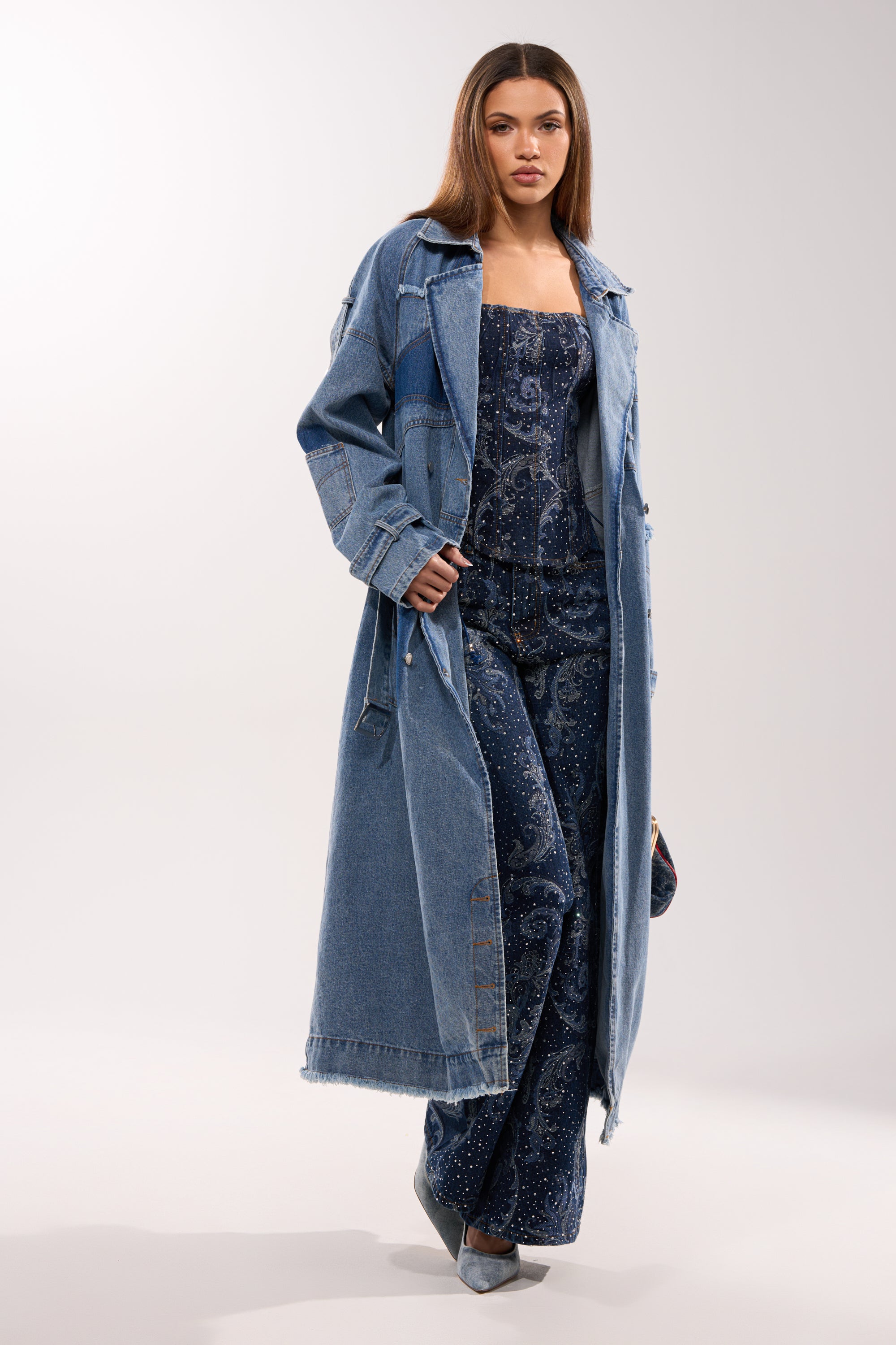 A woman models the RILEY PATCHWORK DENIM TRENCH with a longline silhouette over a dark blue patterned jumpsuit and matching pointed-toe heels, posed against a plain white background. 4