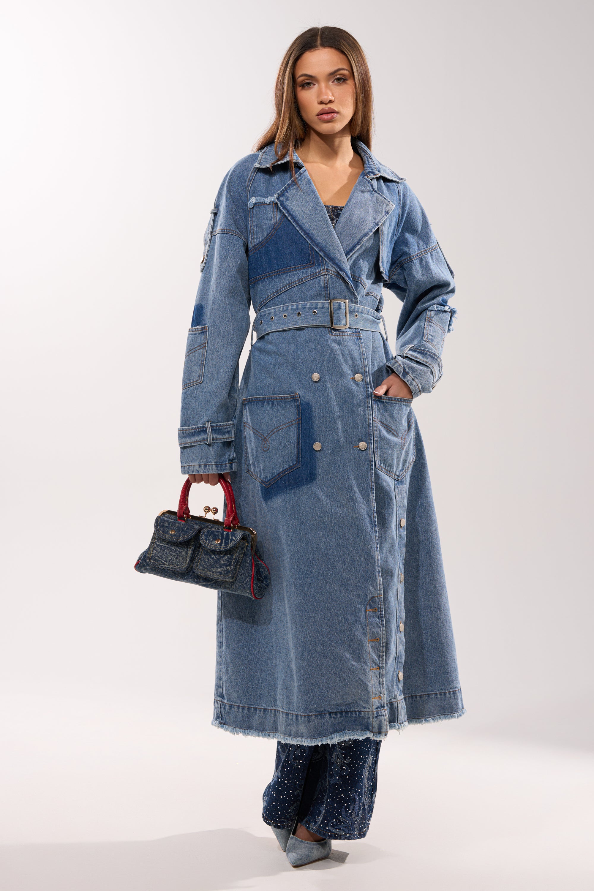 A woman models the RILEY PATCHWORK DENIM TRENCH, featuring a longline vintage-wash silhouette and patchwork details. She pairs it with glittery blue pants, pointed shoes, and a small handbag with red handles, looking confidently at the camera. 5