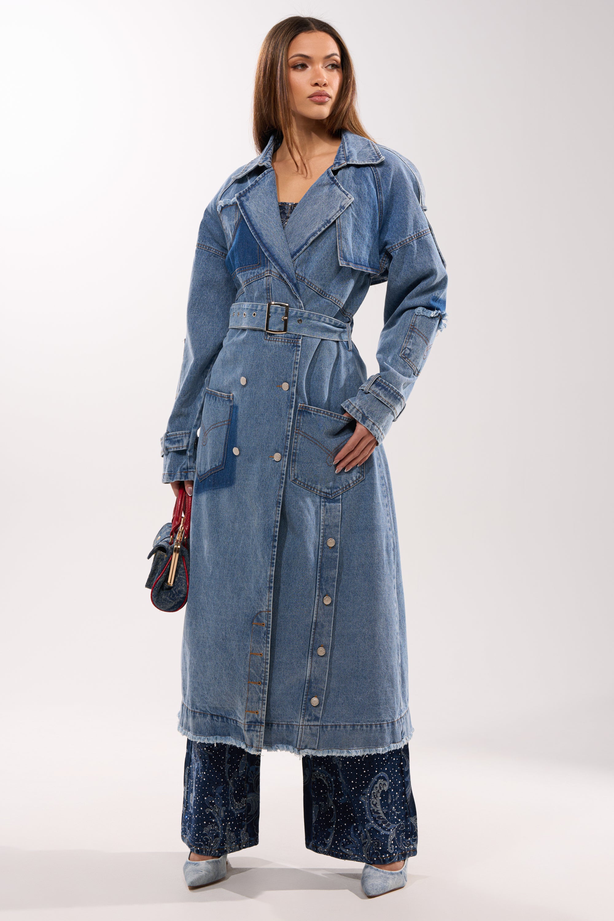 A woman wears the RILEY PATCHWORK DENIM TRENCH, a longline vintage-wash coat with patch pockets and snap buttons, paired with wide-leg dark sparkly pants, pointed heels, and a small handbag. 6