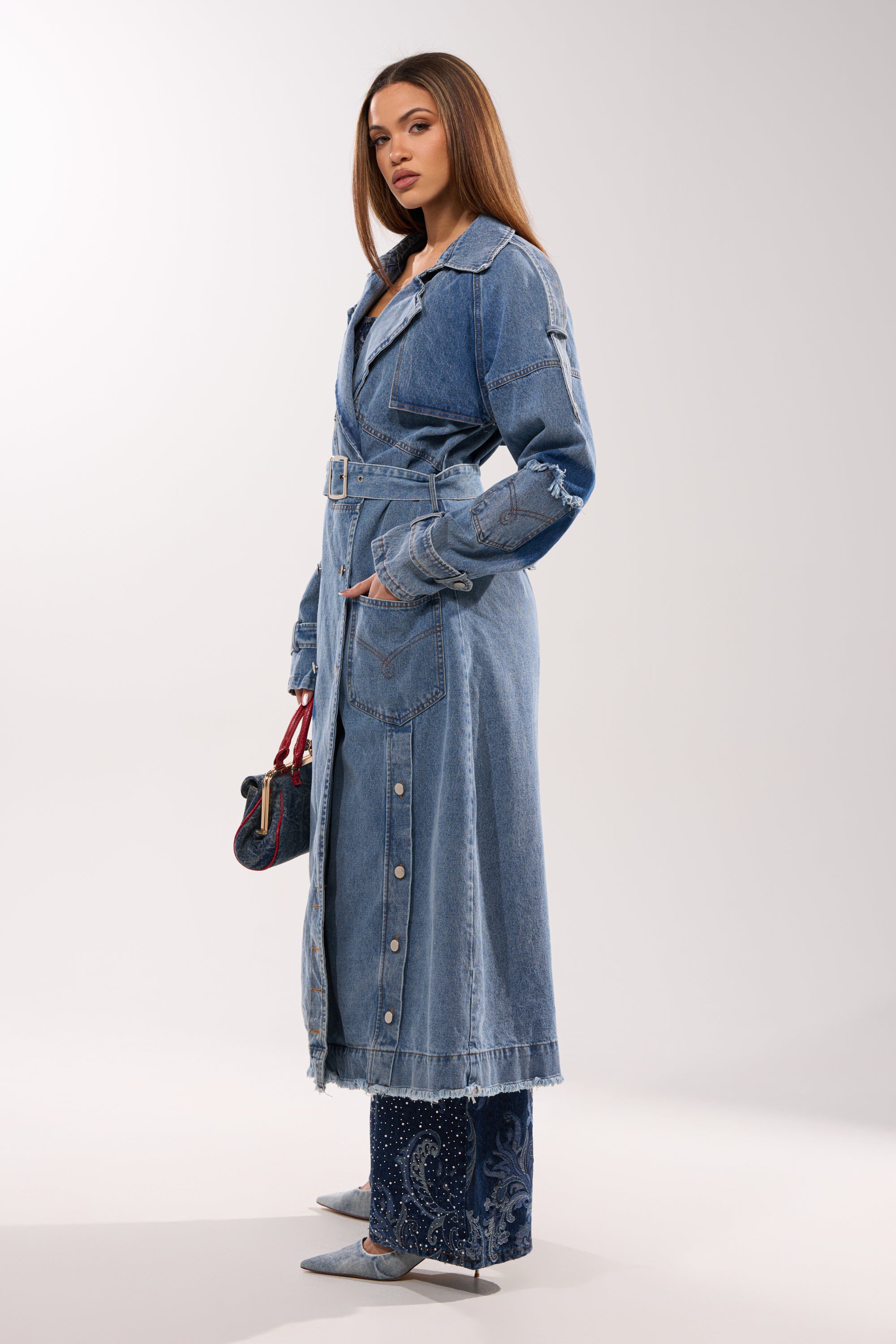 A woman wears the RILEY PATCHWORK DENIM TRENCH, styled with blue patterned pants, pointed gray heels, and a small handbag with a red handle, looking confidently to the side. 7