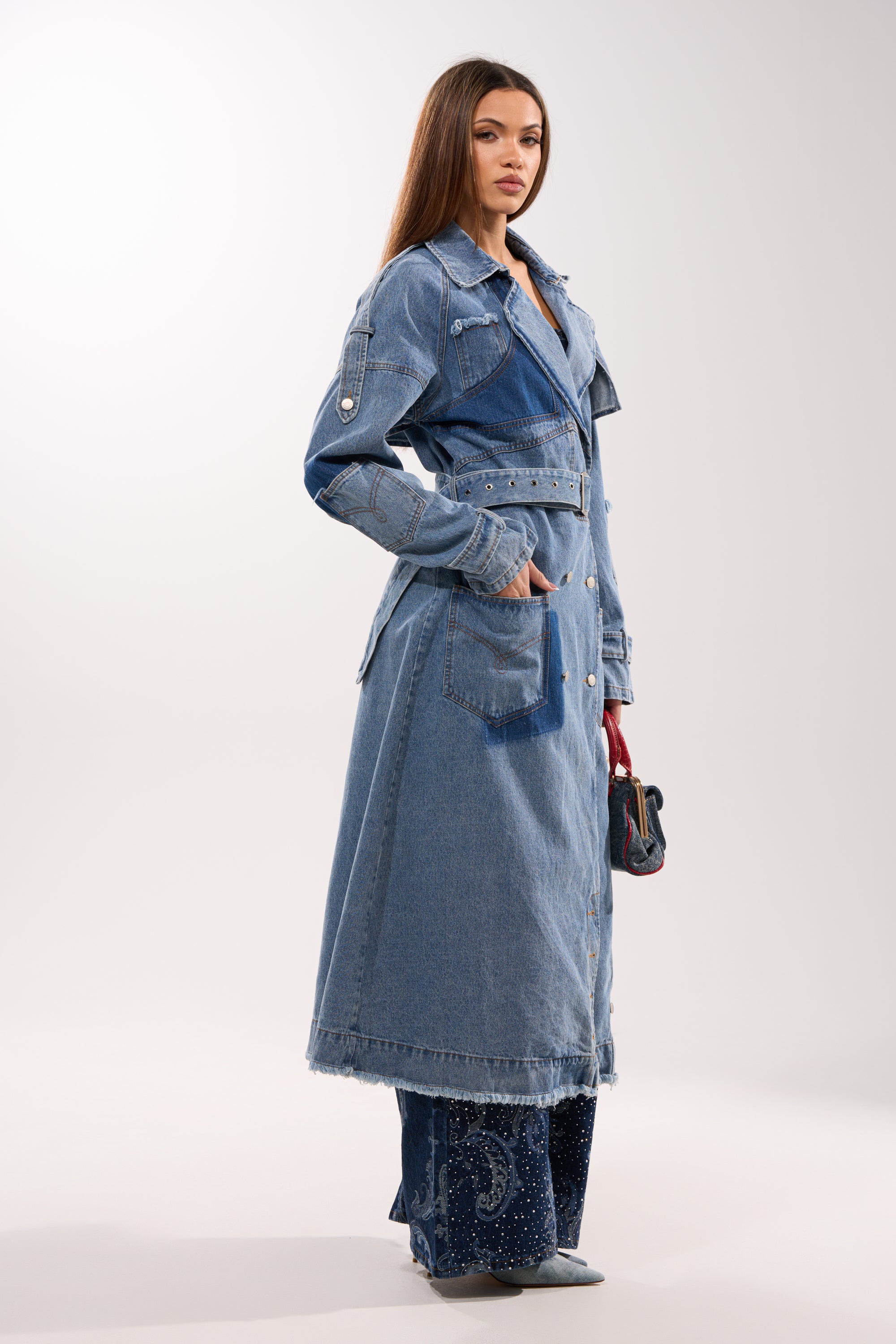 A woman stands confidently in the RILEY PATCHWORK DENIM TRENCH, a vintage-wash, longline denim coat, layered over flared embellished jeans. She holds a small handbag and poses against a plain light background with straight hair. 8