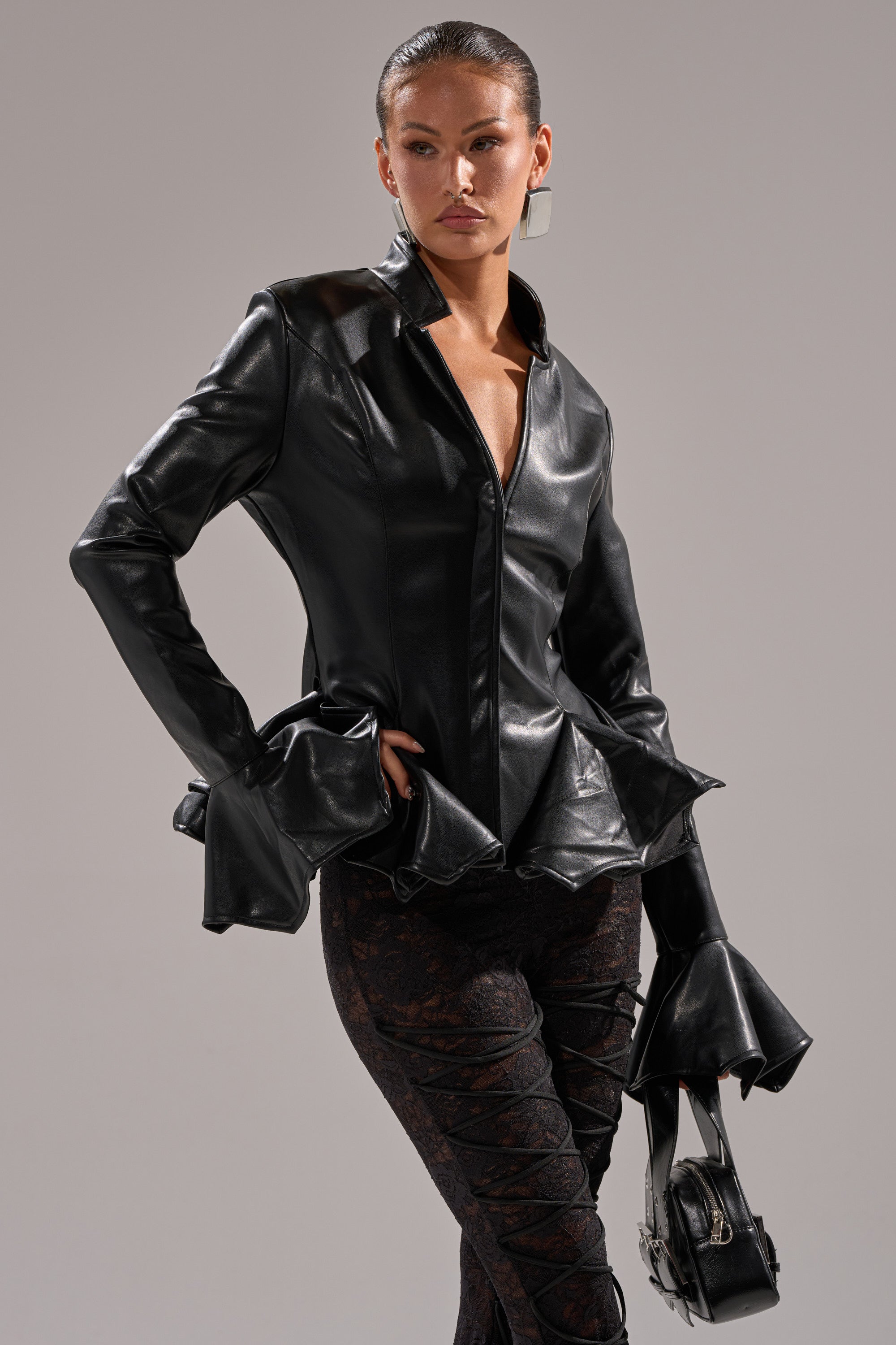 A woman models the CARMEN BODYCON BLAZER IN BLACK, featuring dramatic ruffled cuffs and collar, styled with patterned black pants and a small black handbag. She wears her hair slicked back and looks confident.