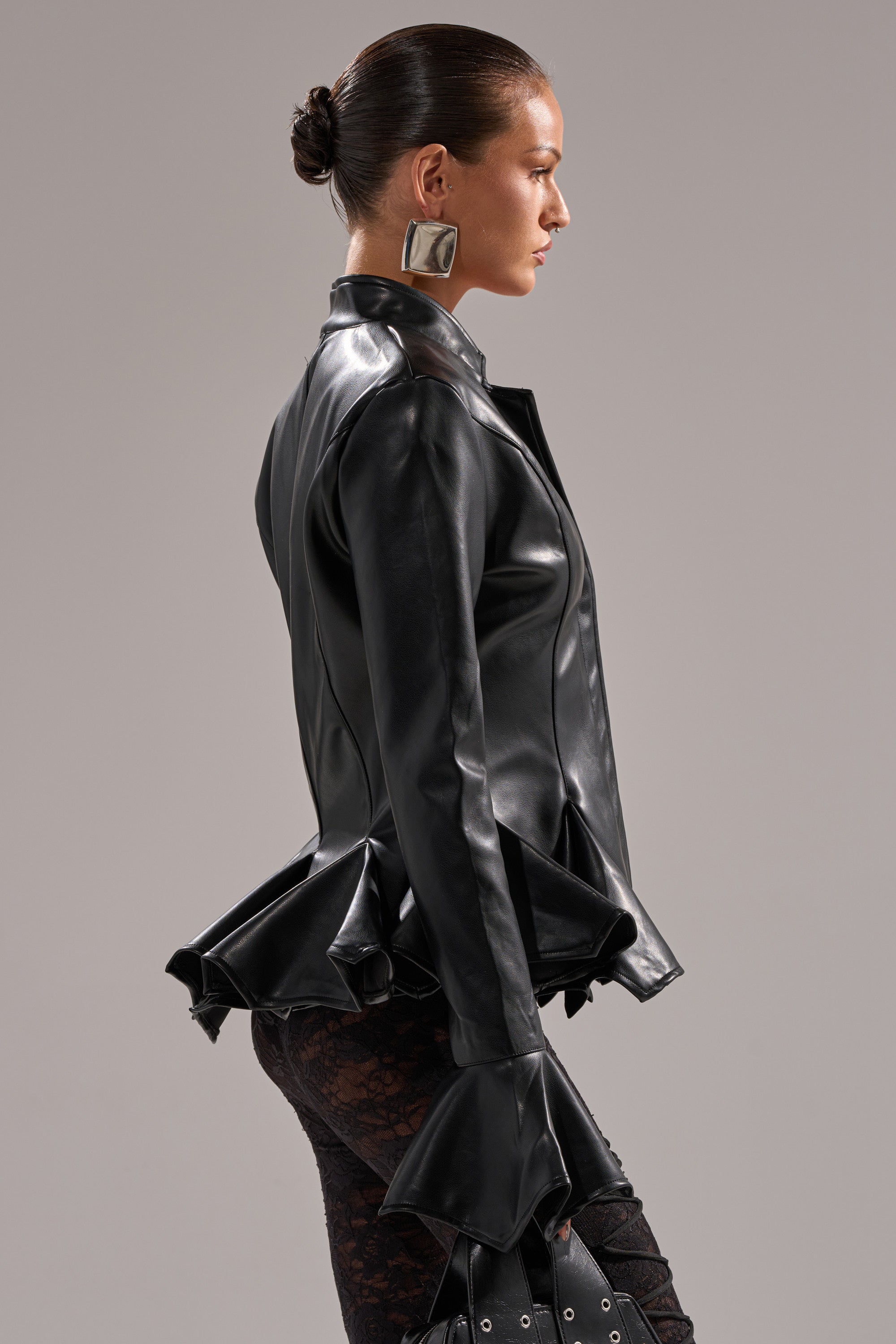 A woman with slicked-back hair models the CARMEN BODYCON BLAZER IN BLACK, featuring a peplum silhouette and flared cuffs, styled with bold square earrings and lace-patterned pants, posing in profile against a plain gray background.