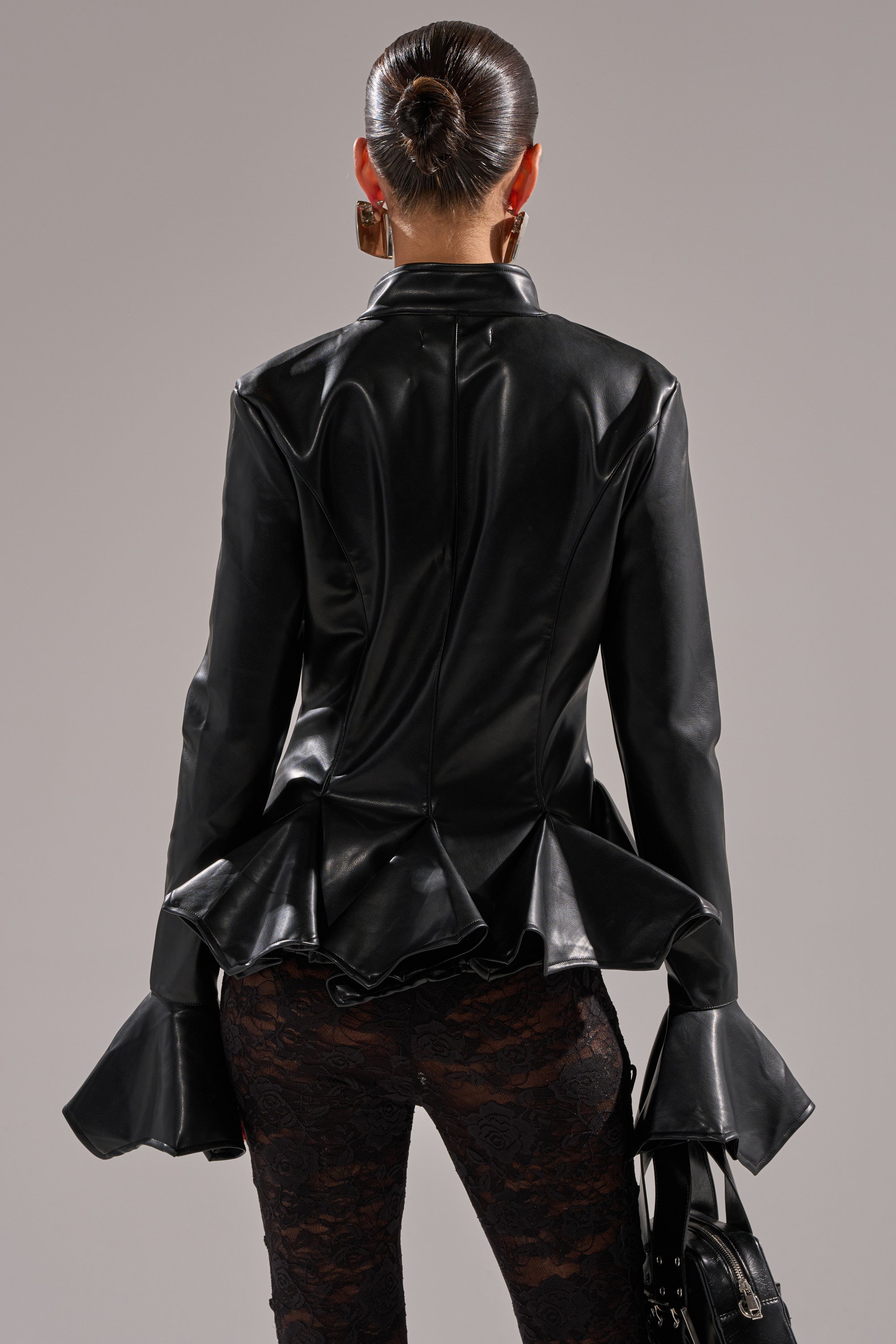 A person with dark hair in a sleek bun wears the CARMEN BODYCON BLAZER IN BLACK, featuring ruffled cuffs and hem, paired with black lace pants against a plain gray background.