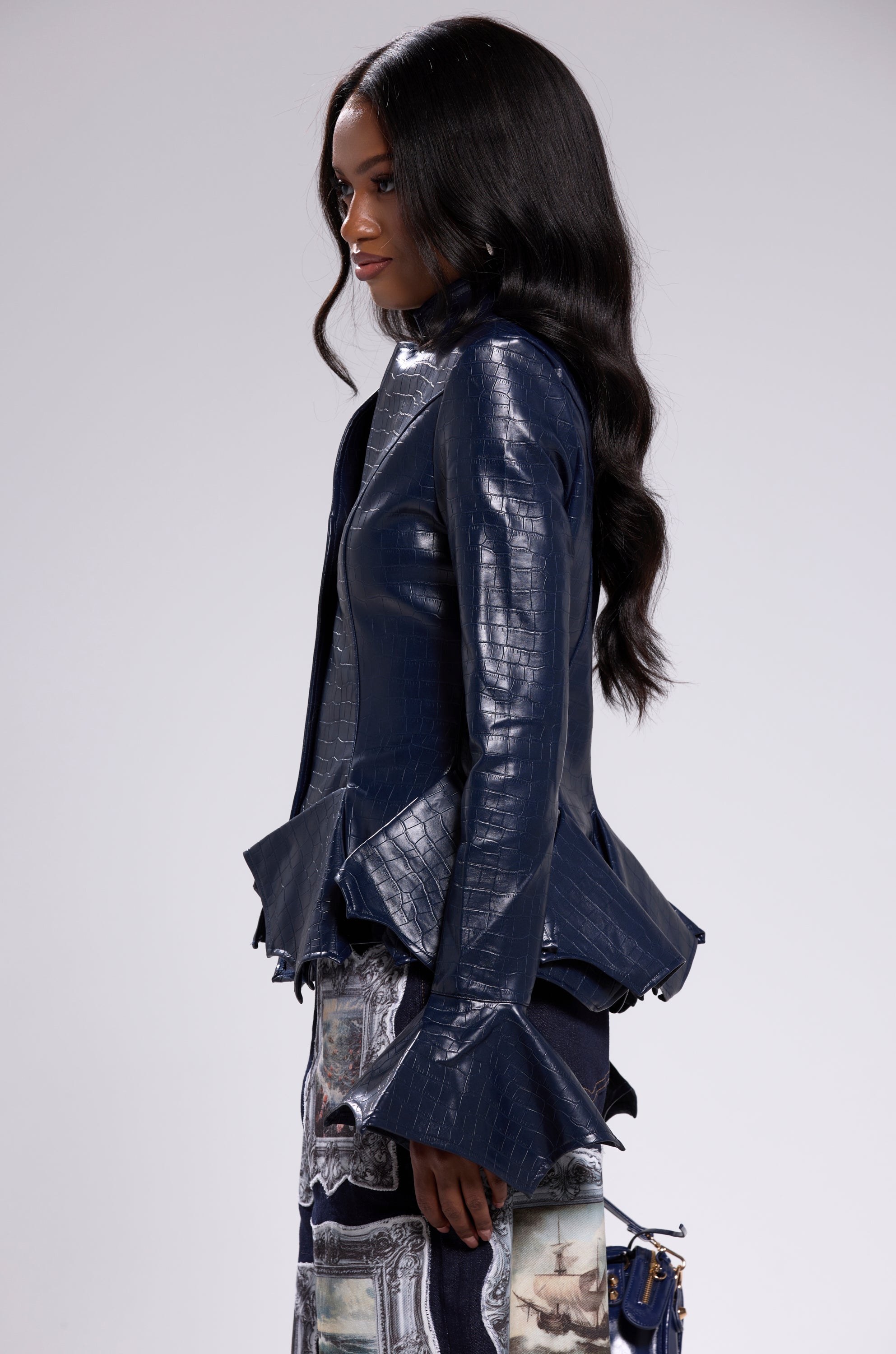 A woman with long wavy hair wears the CARMEN BODYCON BLAZER IN NAVY, featuring a fitted silhouette with flared cuffs and hem, paired with a printed skirt. She stands in profile against a light gray background.