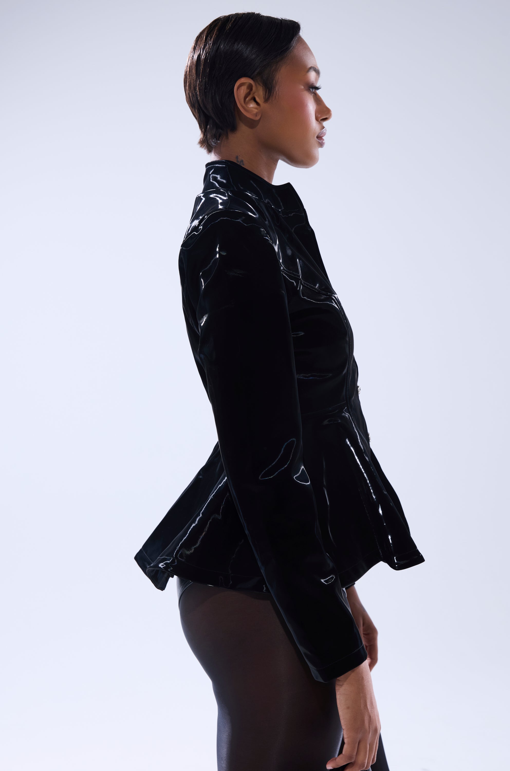 A person in profile against a light background wears the SLIMM LIQUID VELVET BLAZER and sheer black tights, sporting short, sleek hair. 3