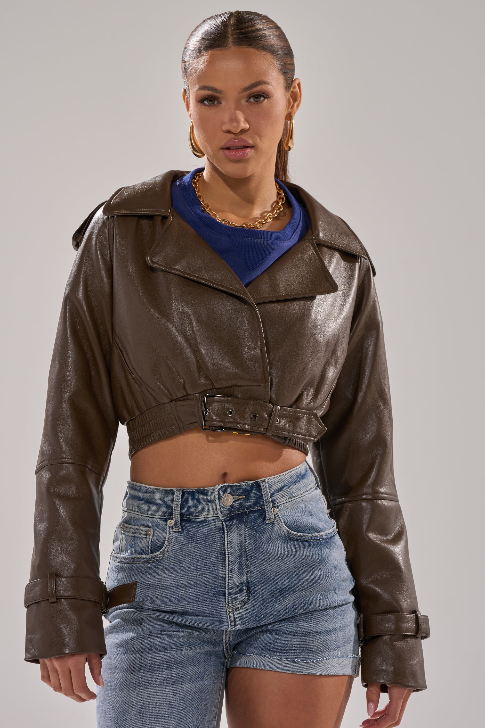 A woman stands against a neutral background wearing the BUTTER LUXE MOTO IN CHOCOLATE, styled with a blue top, gold necklace, hoop earrings, and denim shorts. 0