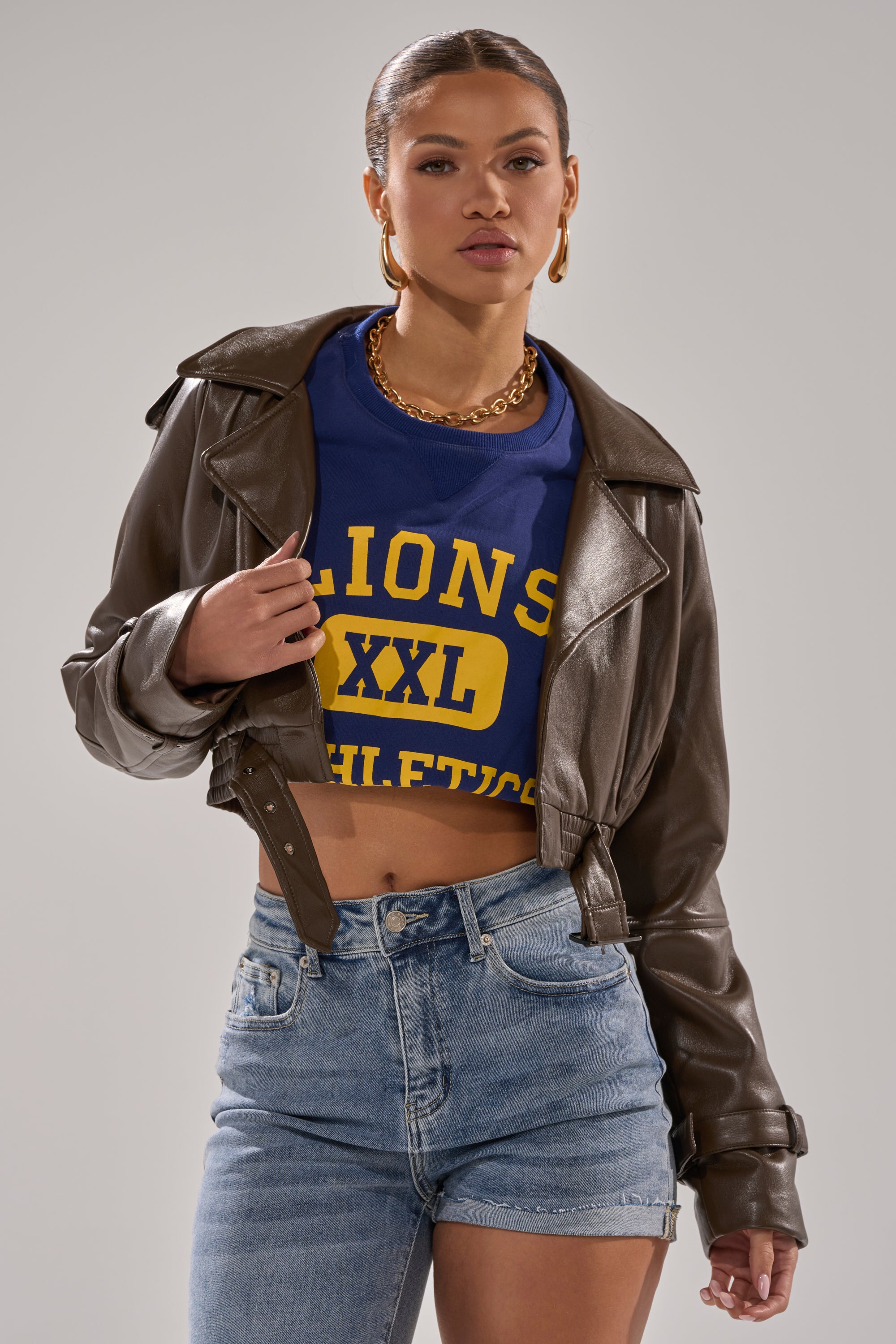 A woman stands confidently against a neutral background in the BUTTER LUXE MOTO IN CHOCOLATE jacket over a blue and yellow "LIONS XXL ATHLETICS" T-shirt and high-waisted denim shorts. 1