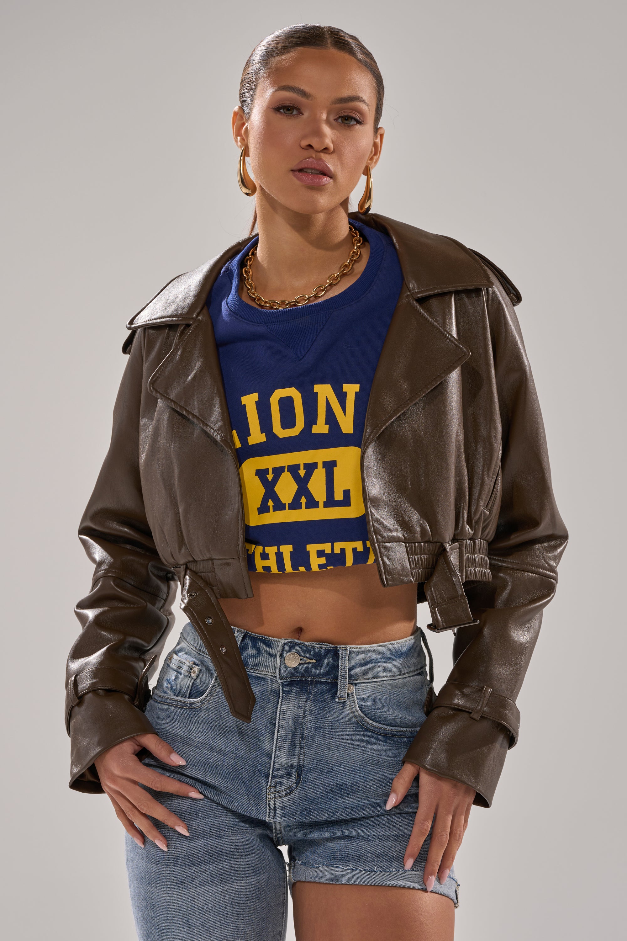 A woman wears the BUTTER LUXE MOTO IN CHOCOLATE, a cropped faux leather jacket, over a blue and yellow graphic crop top with high-waisted denim shorts, hoop earrings, a gold chain necklace, and her hair pulled back. 2