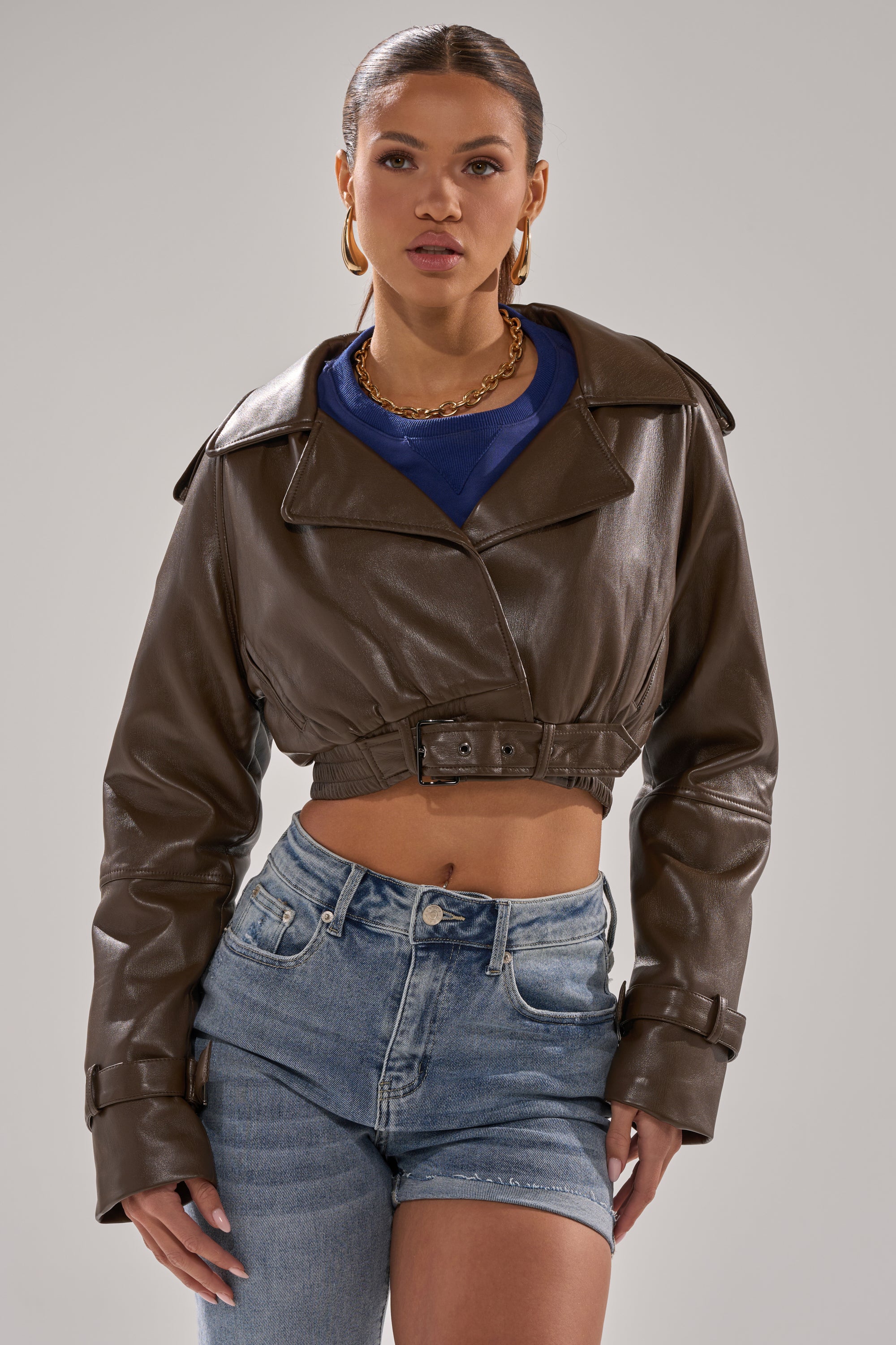 A woman wearing the BUTTER LUXE MOTO IN CHOCOLATE faux leather jacket, styled cropped over a blue top and high-waisted denim shorts, stands against a plain light background, gazing confidently at the camera. 3