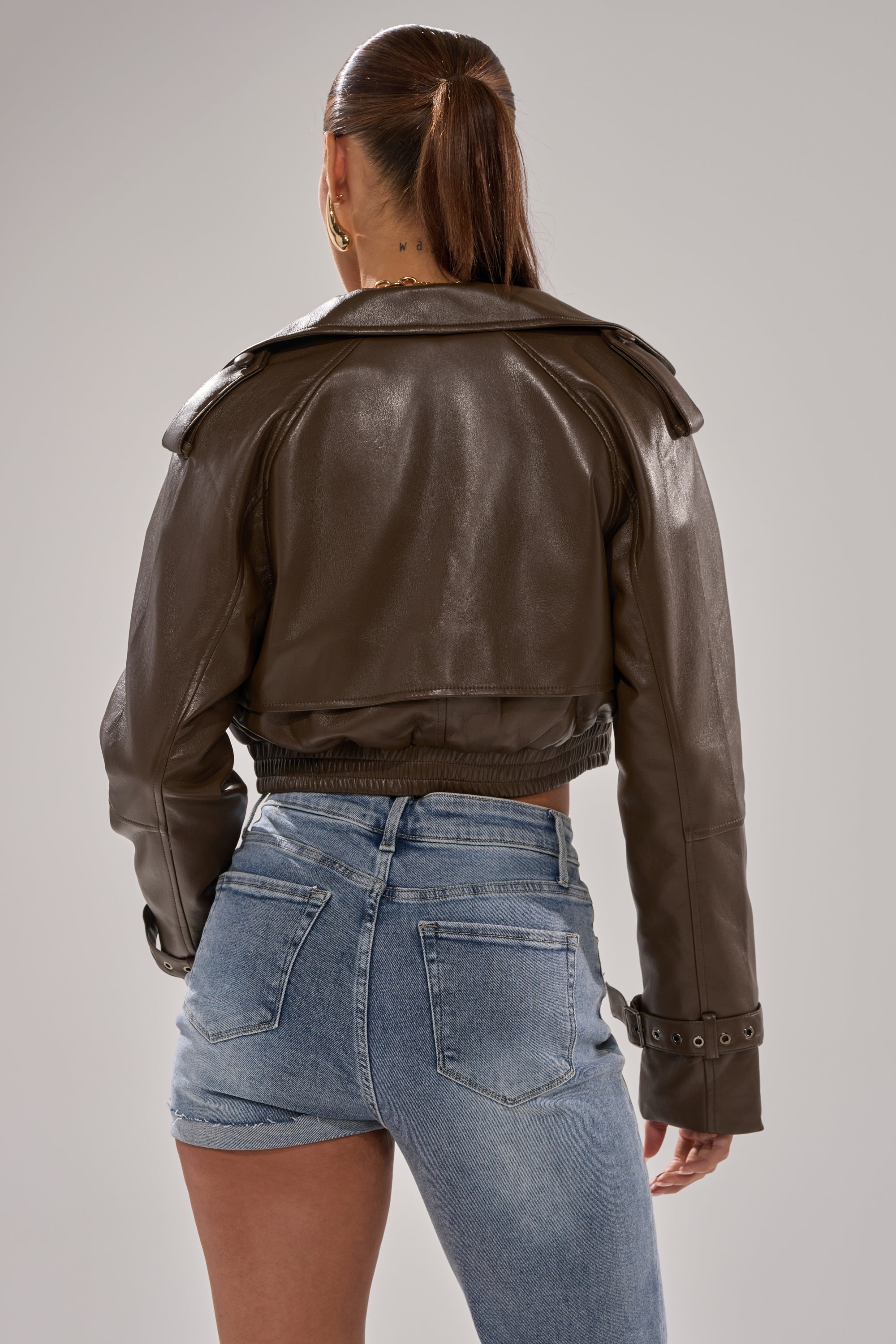 A person with long hair in a ponytail wears the BUTTER LUXE MOTO IN CHOCOLATE jacket and high-waisted blue denim shorts, standing with their back to the camera against a neutral background. 5