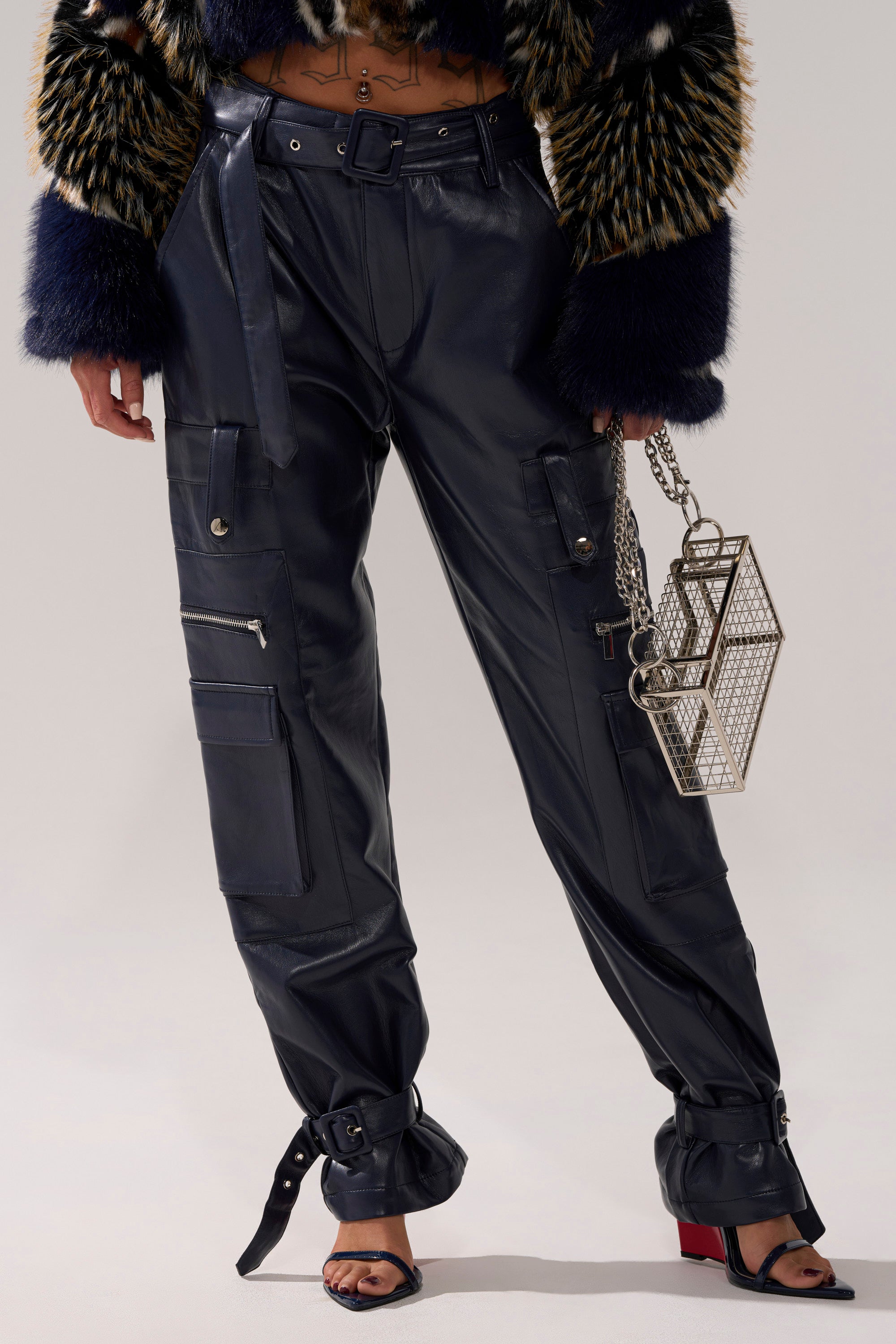 A person wears high-waisted navy BUTTER LUXE PANTS with ankle straps, a patterned faux fur jacket, black heeled sandals, and holds a geometric silver mesh handbag against a plain light-colored background.