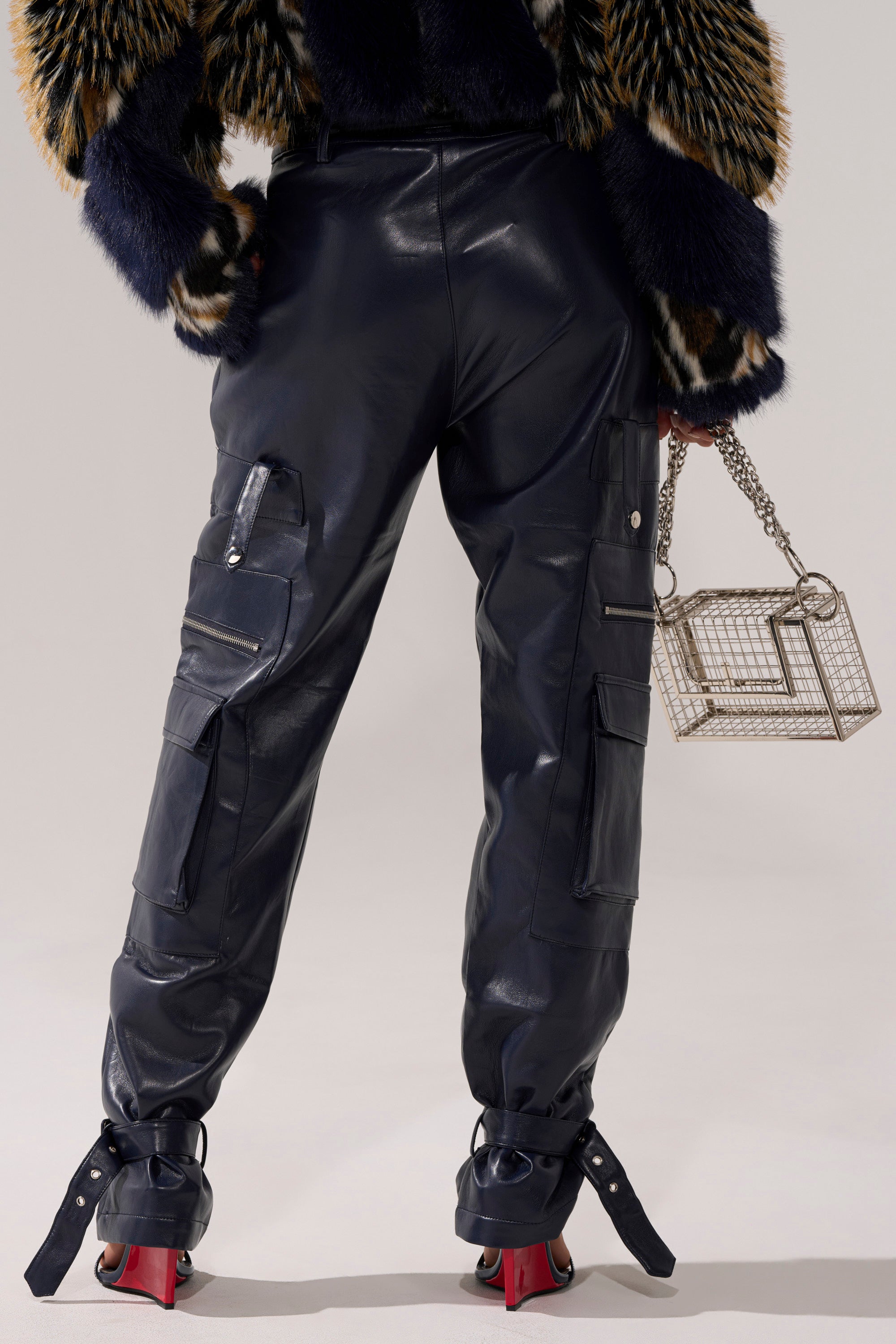 A person stands facing away, wearing BUTTER LUXE PANTS IN NAVY, a fur-patterned jacket, and red-heeled shoes. They hold a metallic cage-style handbag with a chain handle in their right hand.