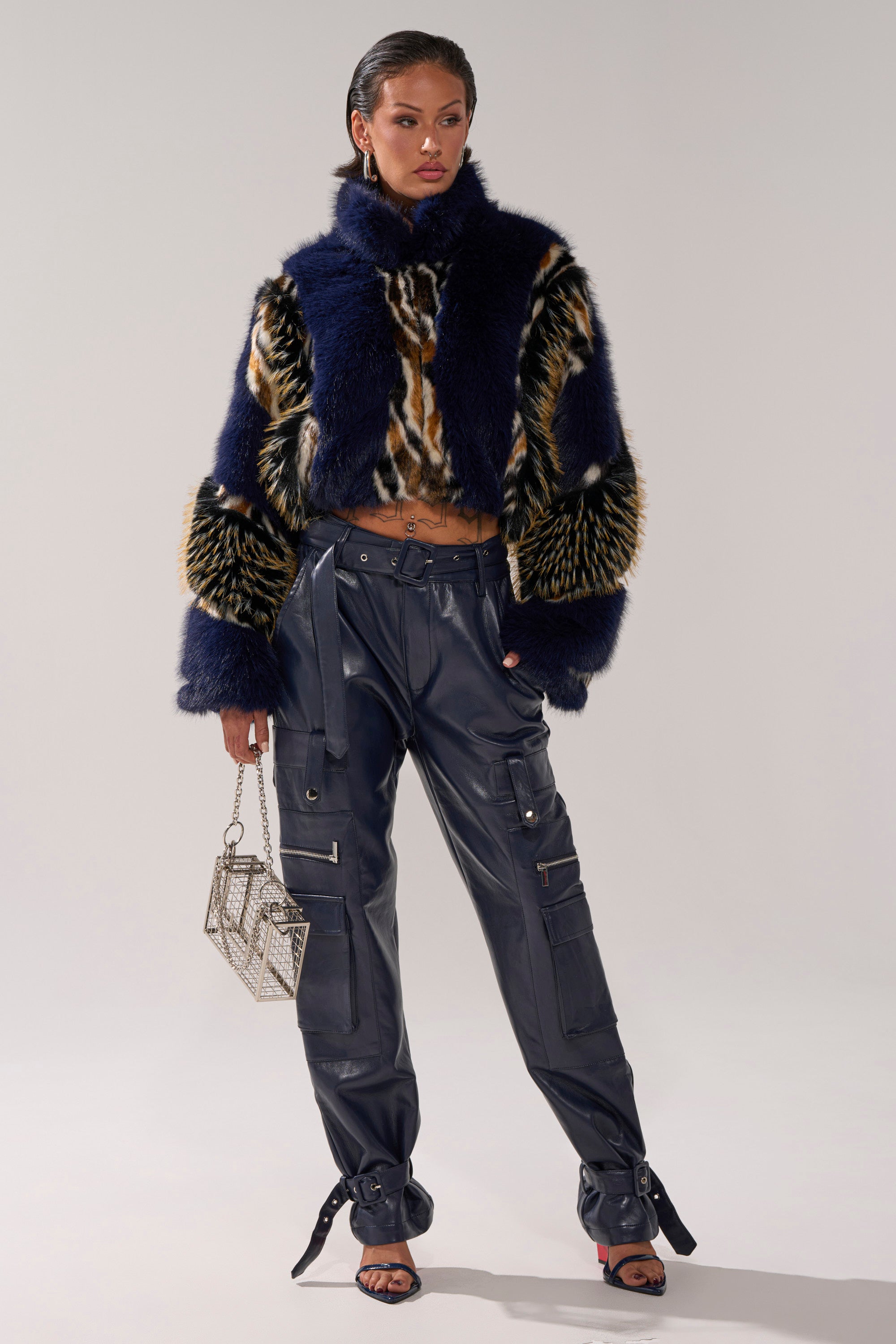 A woman poses in a furry animal print jacket, BUTTER LUXE PANTS IN NAVY with zippers and straps, open-toe heels with bows, and holds a small sparkling handbag.