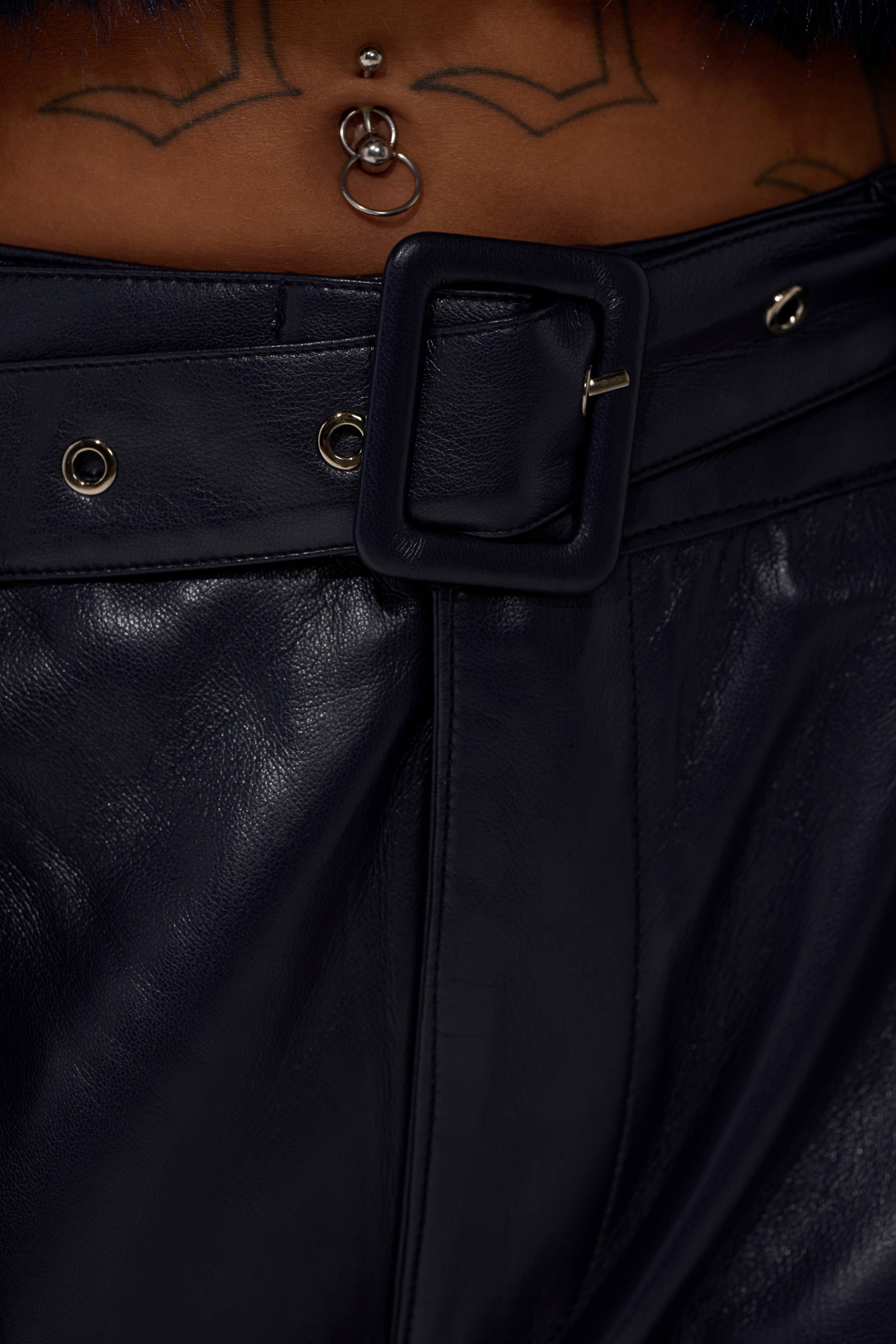 Close-up of a person wearing BUTTER LUXE PANTS IN NAVY, styled high-waisted with a wide belt and large buckle. A body piercing and geometric tattoo on their lower abdomen are also visible.