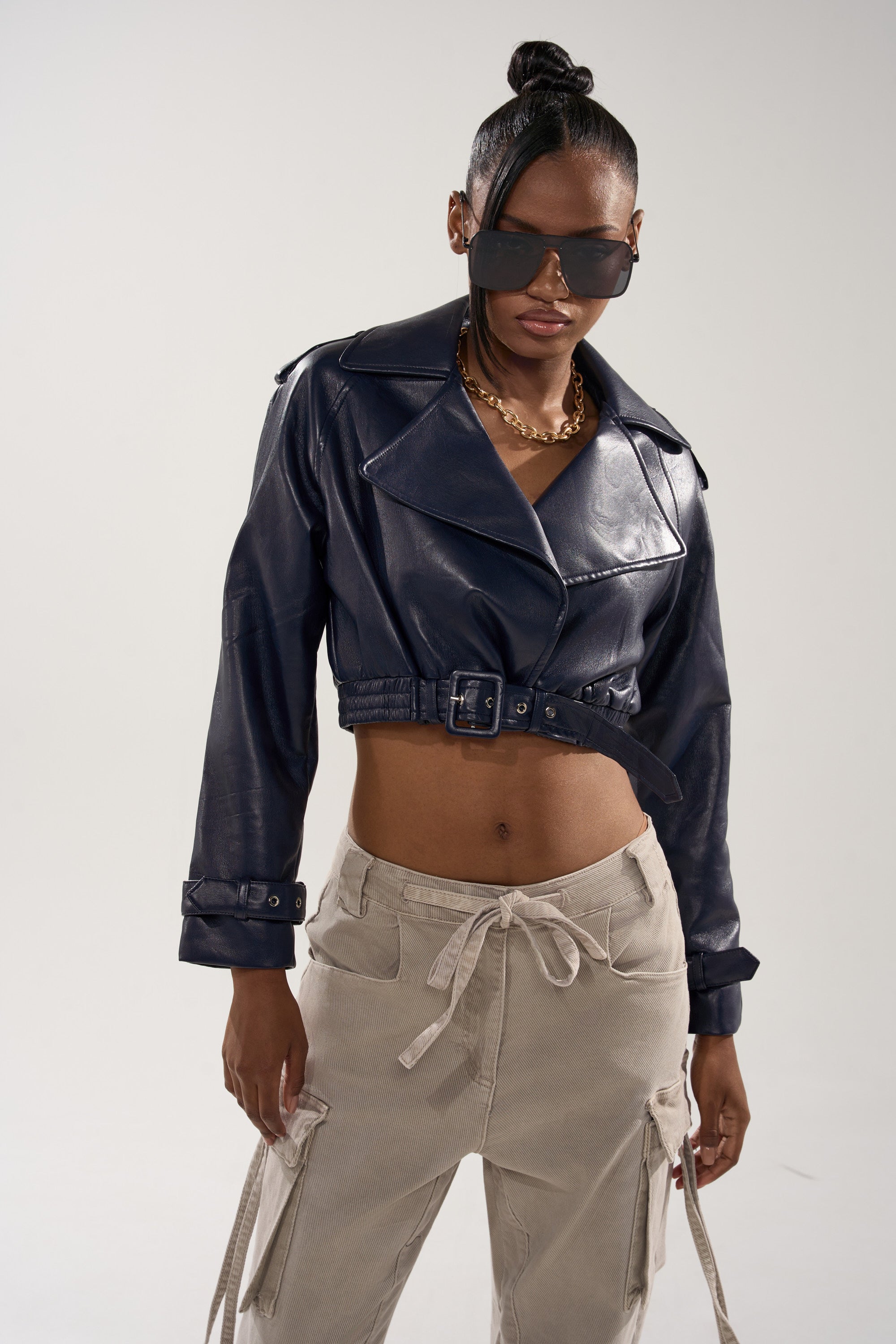 A woman wears the BUTTER LUXE MOTO IN NAVY jacket, beige drawstring cargo pants, sunglasses, and a gold chain necklace while standing against a plain light background. 3