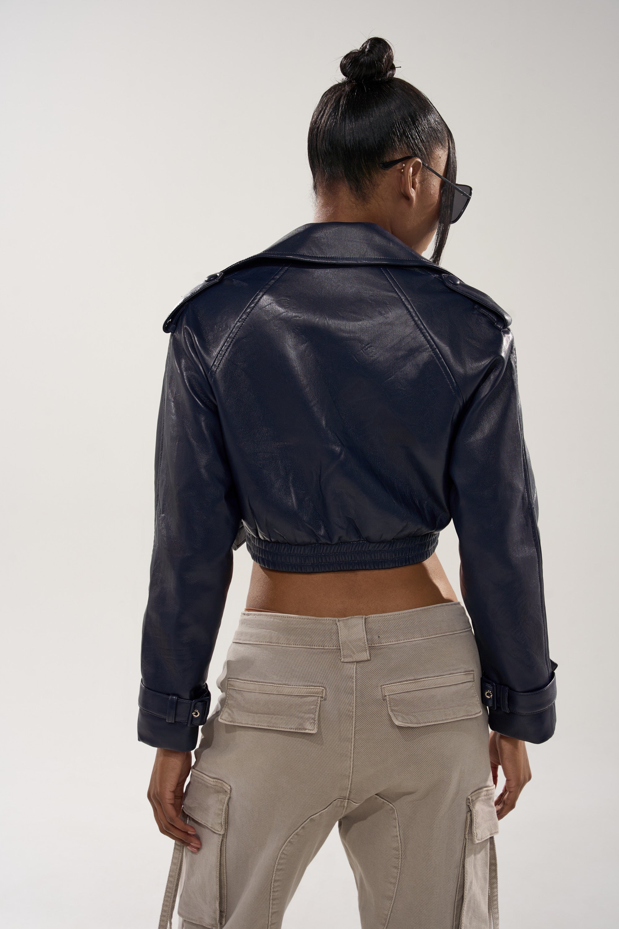 A person with a bun hairstyle and sunglasses is shown from the back against a plain light background, wearing the BUTTER LUXE MOTO IN NAVY cropped faux leather jacket and light cargo pants. 5