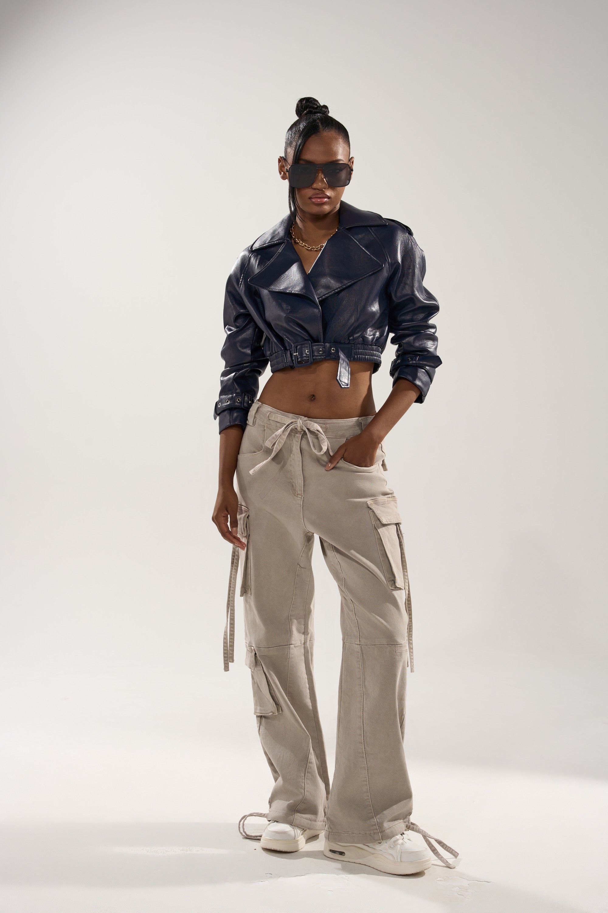 A woman poses confidently in the BUTTER LUXE MOTO IN NAVY, paired with light beige drawstring cargo pants, white sneakers, and sunglasses against a plain light background. 7