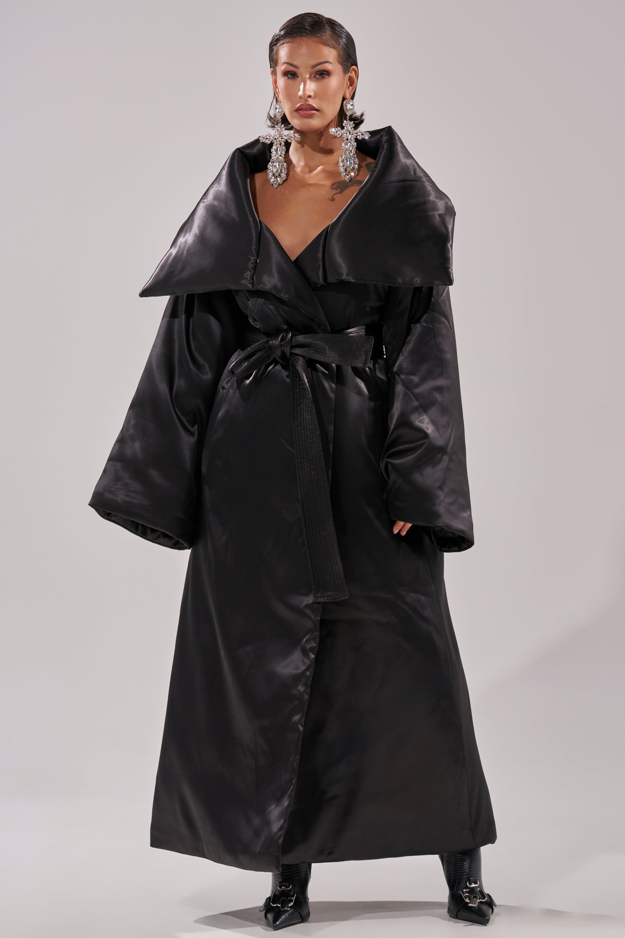 A woman wears THE MOMENT SATIN PUFFER TRENCH, a long black coat with wide lapels and a self-tie waist belt, styled with black boots and sparkling chandelier earrings against a plain light background.