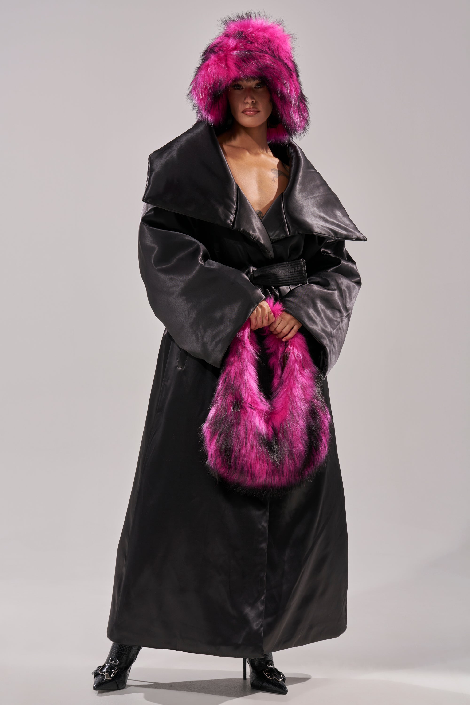 Wearing THE MOMENT SATIN PUFFER TRENCH—a long black coat with an oversized collar and self-tie waist—paired with black high-heeled boots, a large pink-and-black fur hat, and matching handbag, the person stands against a plain light background.