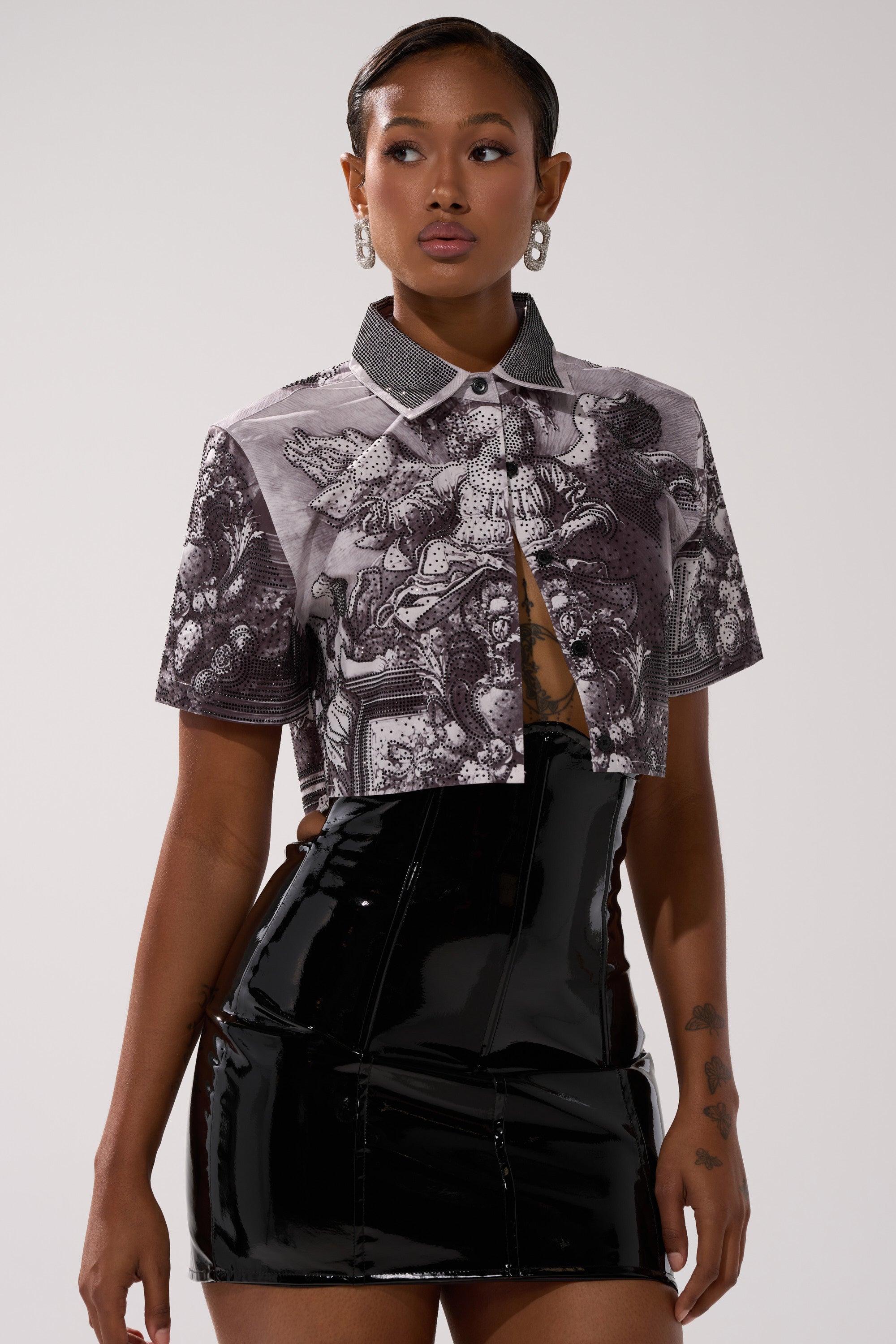 A woman wears the FEELING ALL THE WAVES BUTTON DOWN featuring intricate black-and-white patterns, paired with a shiny black high-waisted skirt. She has short hair, hoop earrings, arm tattoos, and stands against a plain light background.