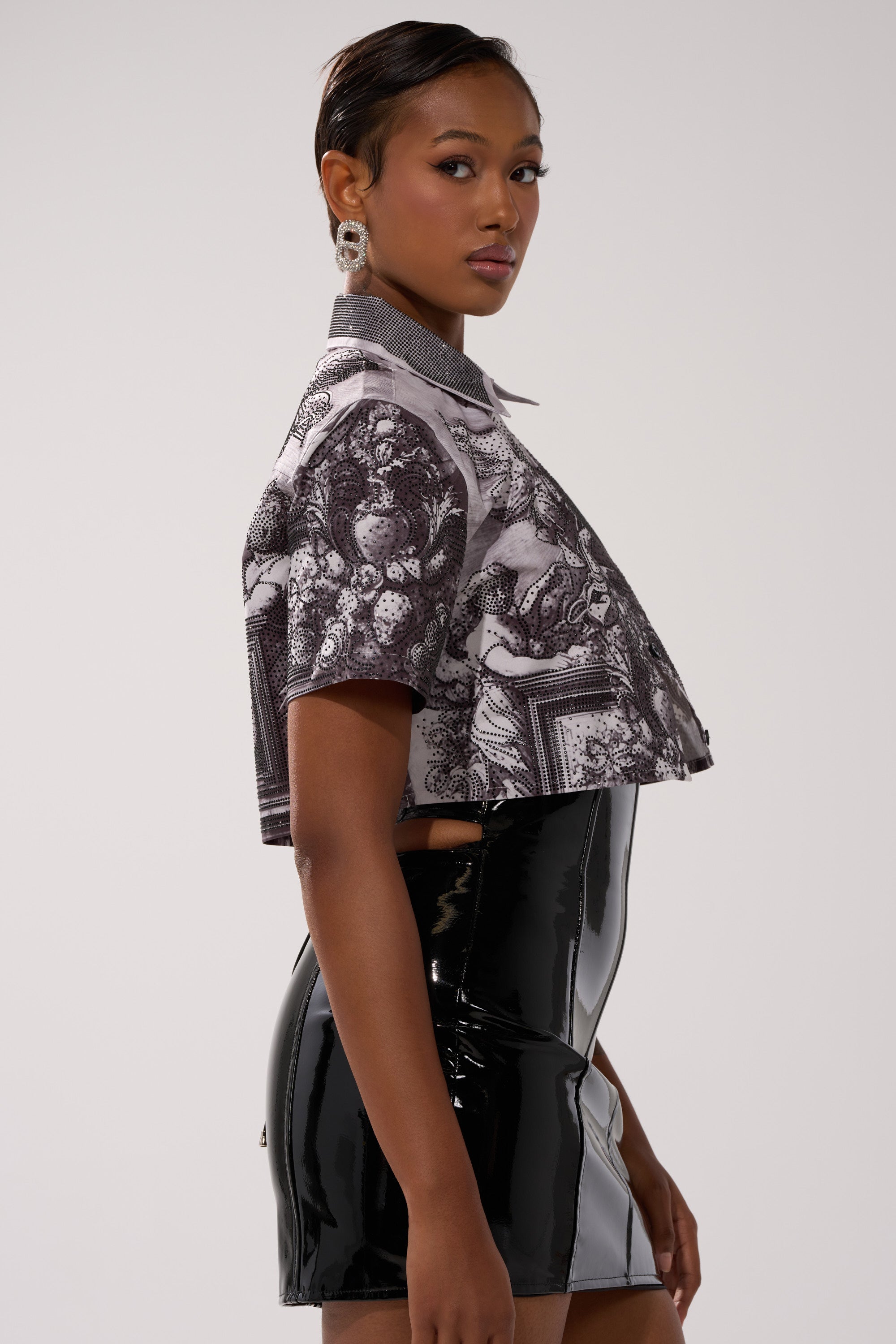 A woman poses in profile against a plain background, wearing the FEELING ALL THE WAVES BUTTON DOWN—a cropped monochrome print shirt—paired with a shiny black high-waisted skirt, statement earrings, and short styled hair.