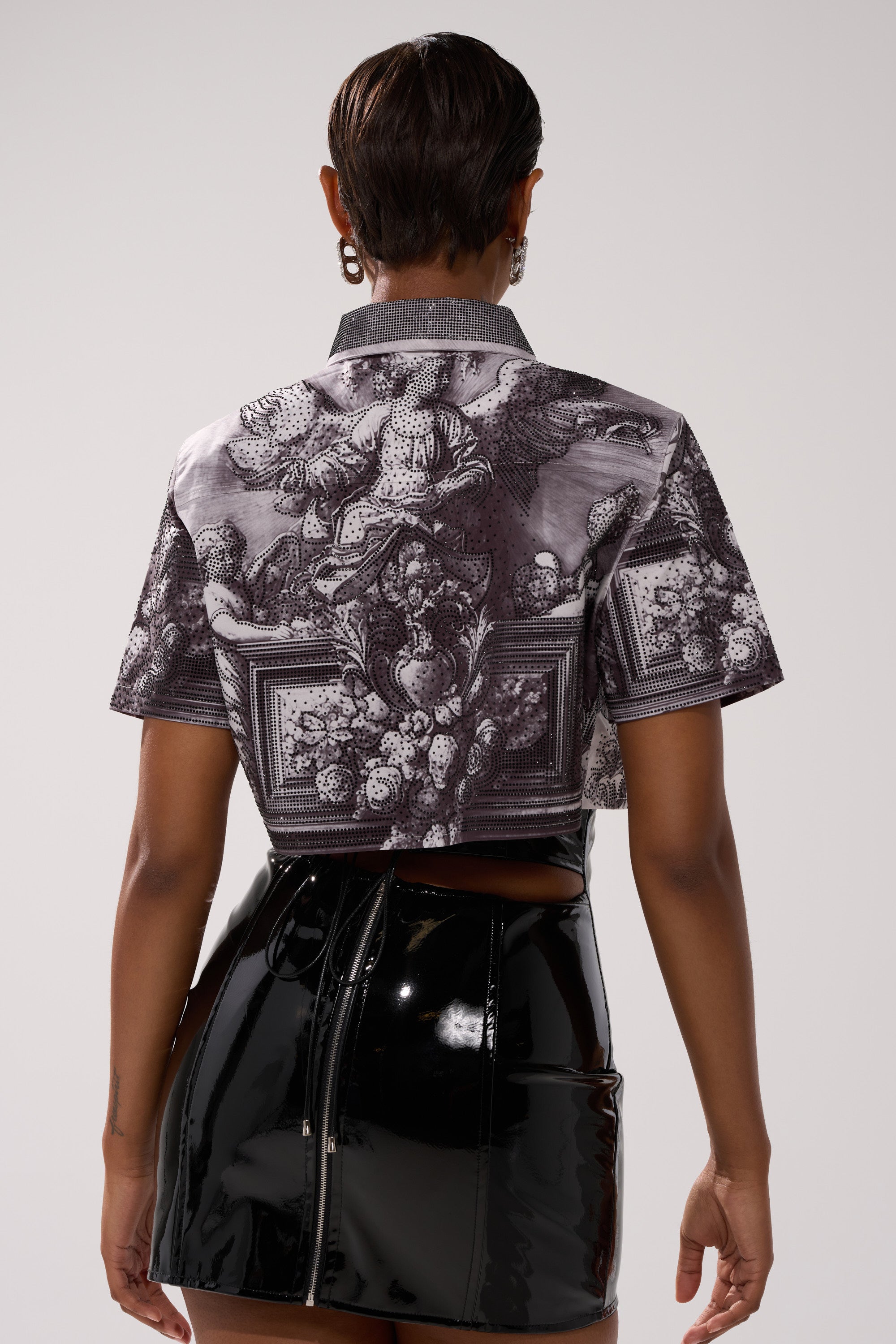 A person stands facing away, wearing the FEELING ALL THE WAVES BUTTON DOWN—a monochrome print, short-sleeve shirt—paired with a shiny black vinyl skirt with a back zipper. Their hair is short and they wear hoop earrings.
