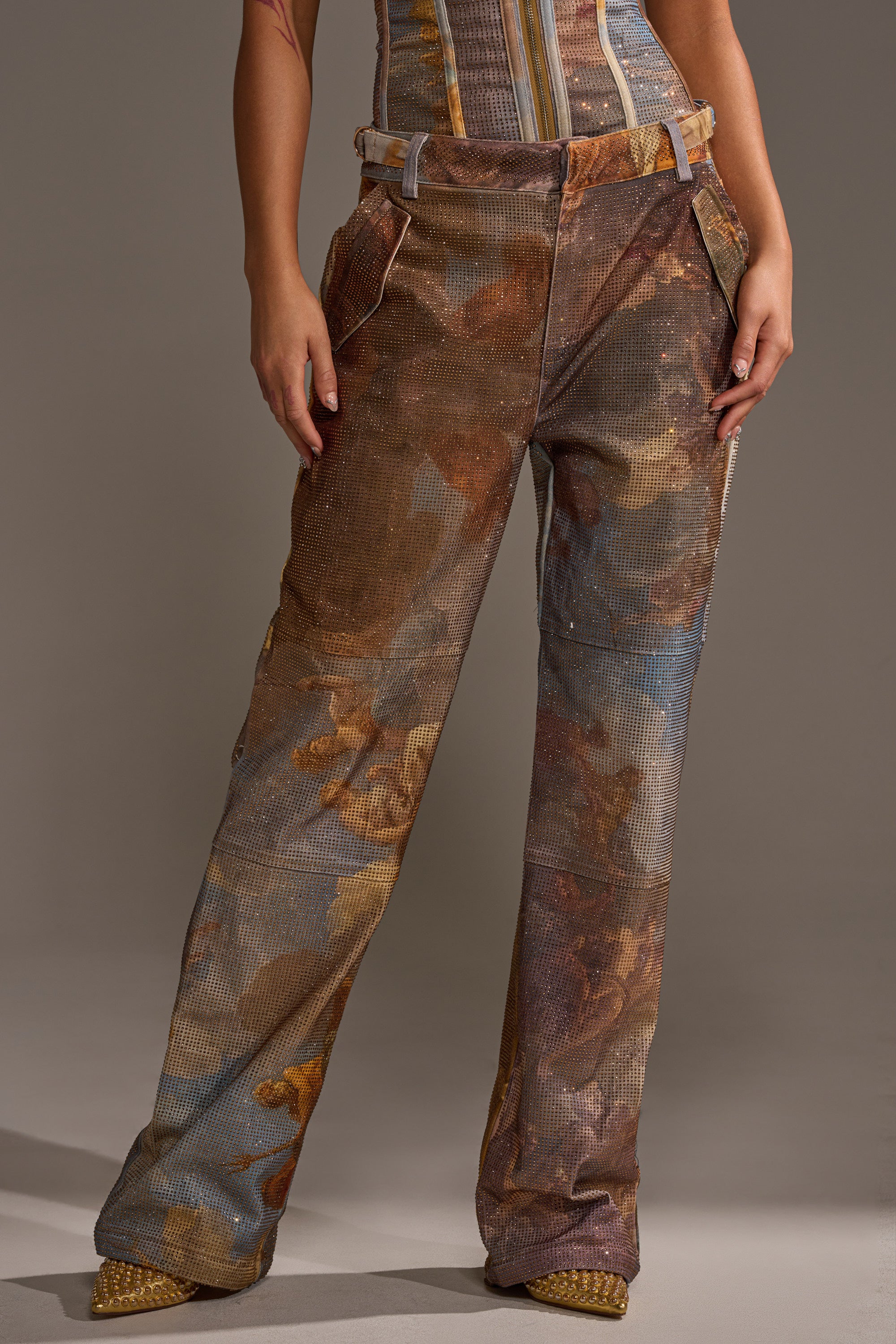 Wearing the IN THE FUTURE TROUSER, a high-waisted, loose-fitting pant with a brown and blue abstract graphic print, paired with a matching corset top and gold pointed heels, the person stands against a plain background.